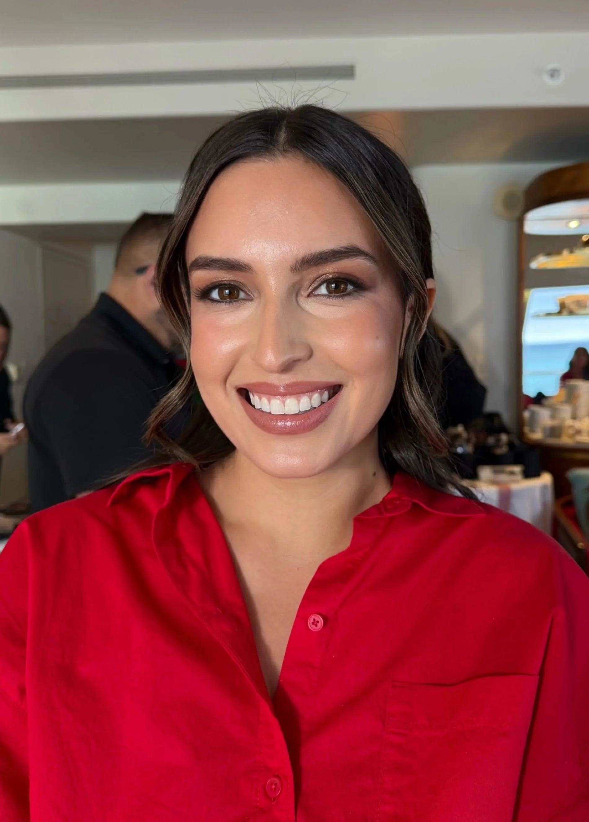 A woman with dark brown hair, brown eyes, and light makeup smiling, wearing a red chef's coat in a restaurant setting with other people and food in the background.