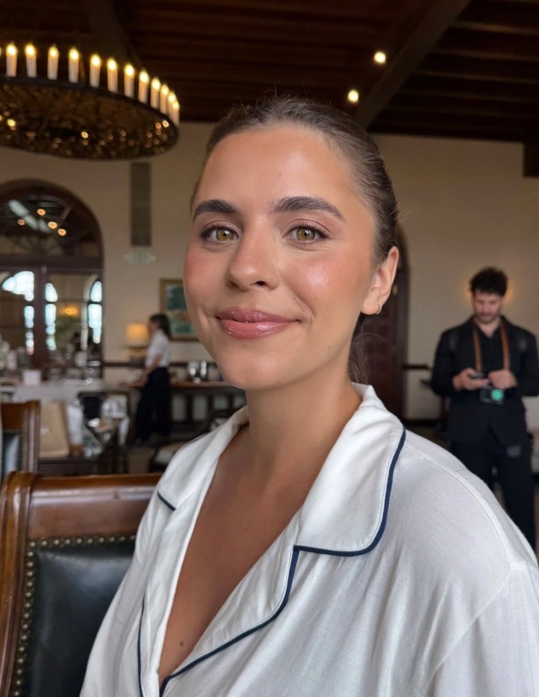 A woman with brown hair tied back, wearing a white silk pajama top with dark piping, smiling at the camera inside a restaurant with wooden ceiling beams, a chandelier, and large windows. A man with a camera is in the background.