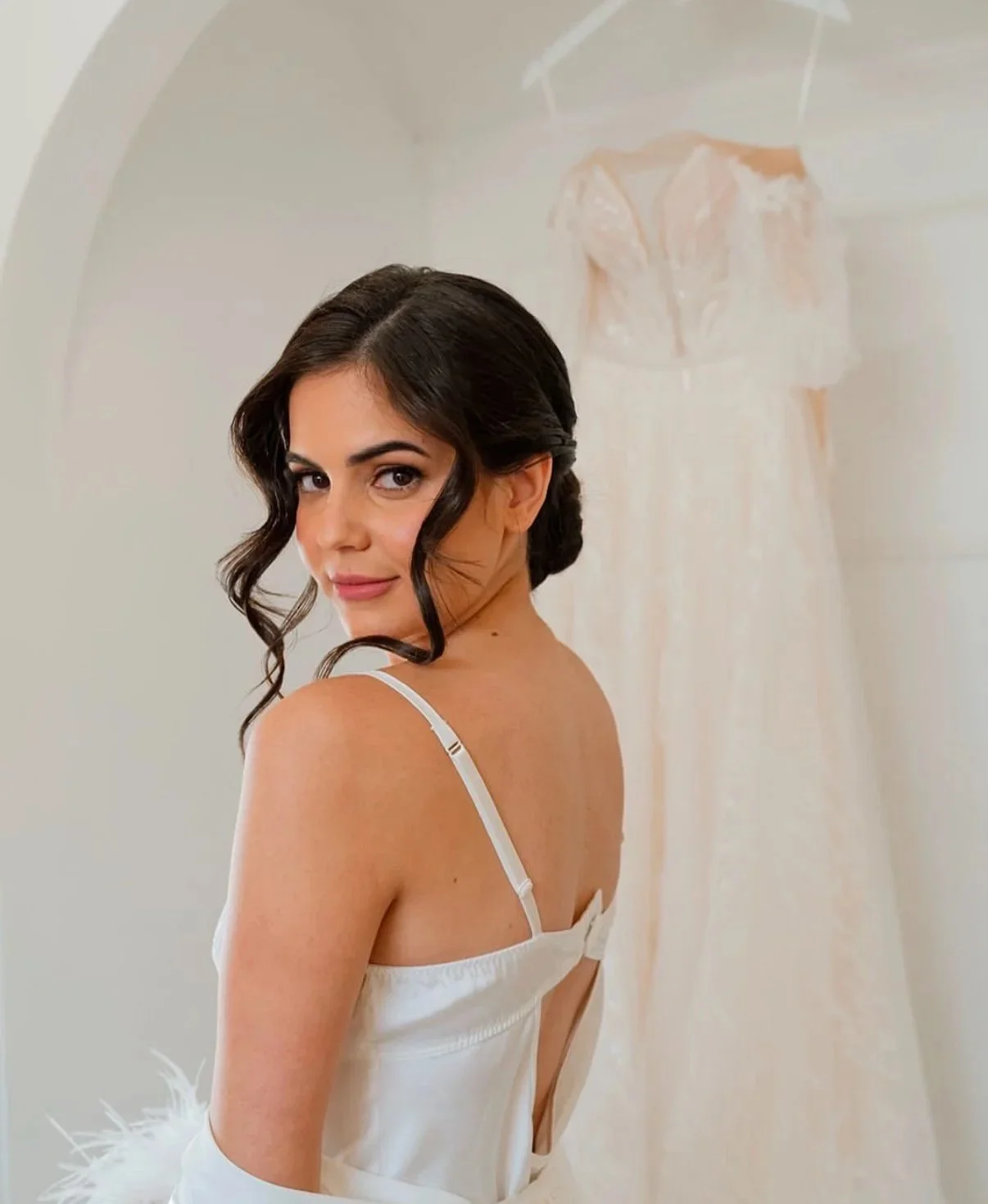 A woman with dark hair styled in loose curls wearing a white satin slip dress, standing in front of a mirror with a wedding dress hanging behind her.