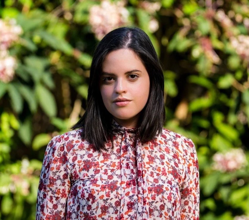 A woman with dark hair and fair skin stands outdoors in front of green shrubs and flowering plants, wearing a floral blouse with a tie at the collar.