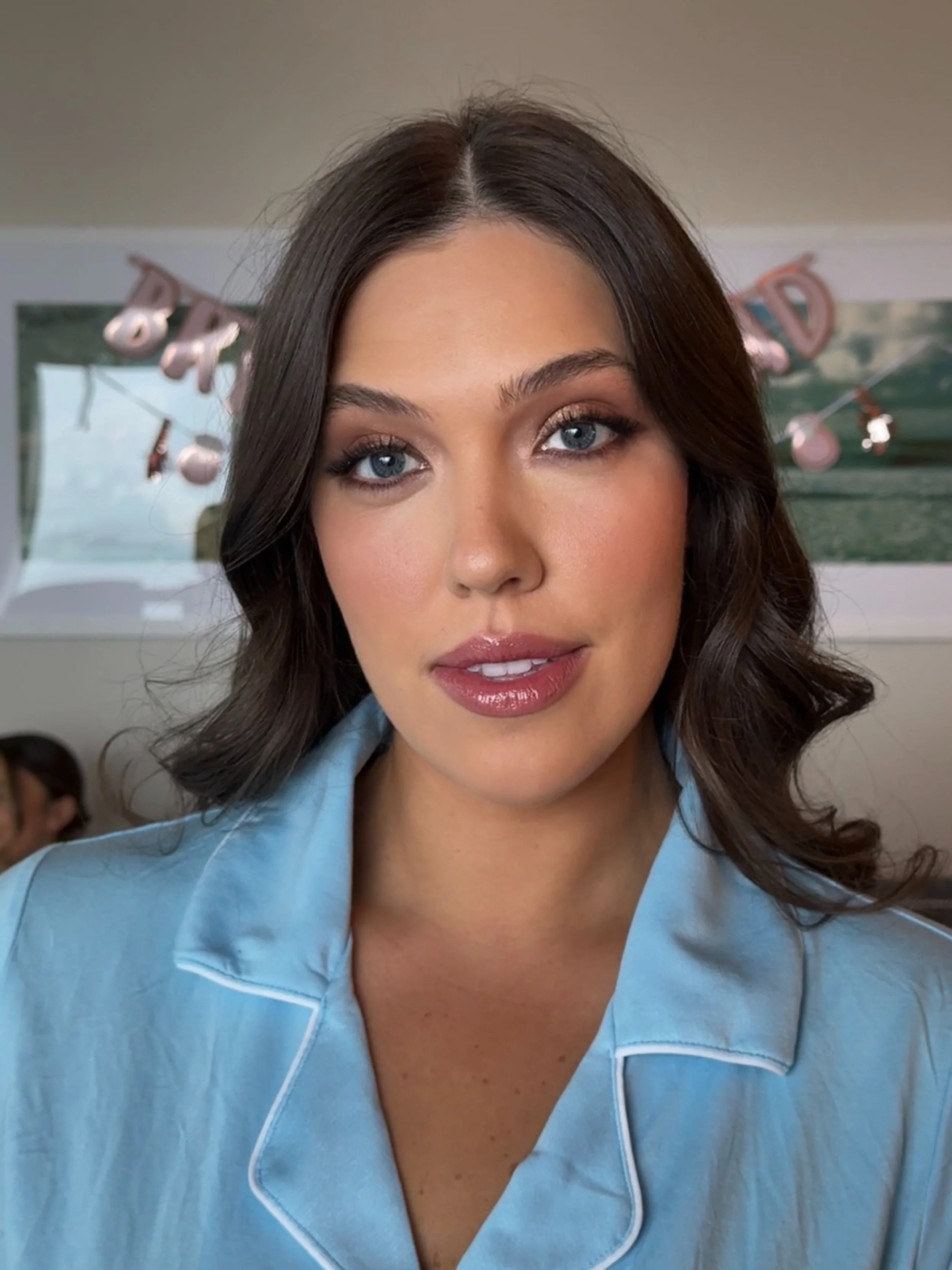 Close-up of a young woman with wavy dark brown hair, wearing light blue pajamas, smiling softly, with a colorful background and a 'Babyshower' banner.