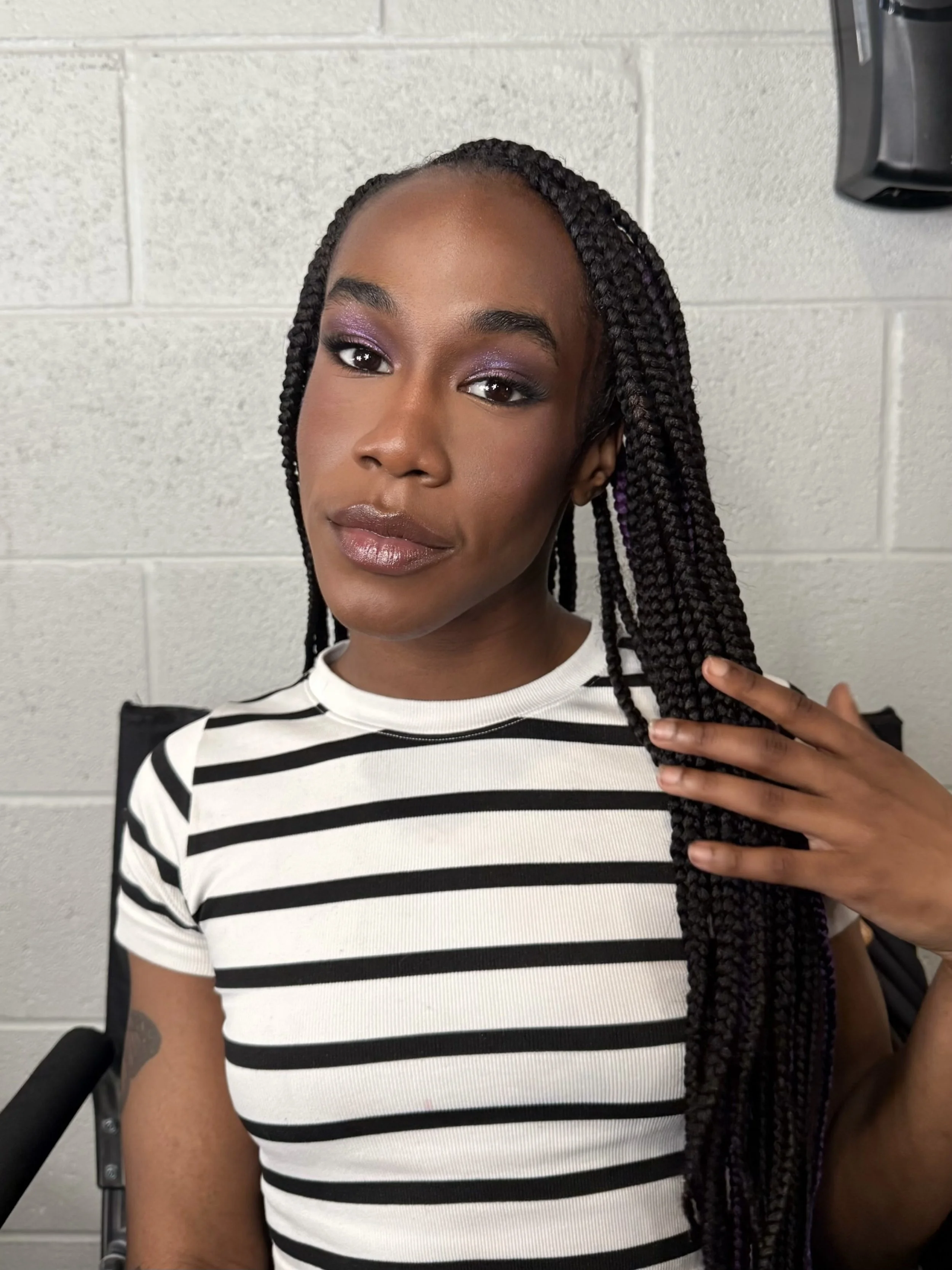 Close-up of a young woman with braided hair, wearing a black and white striped T-shirt, standing against a plain gray wall, holding her braids with one hand, smiling softly.
