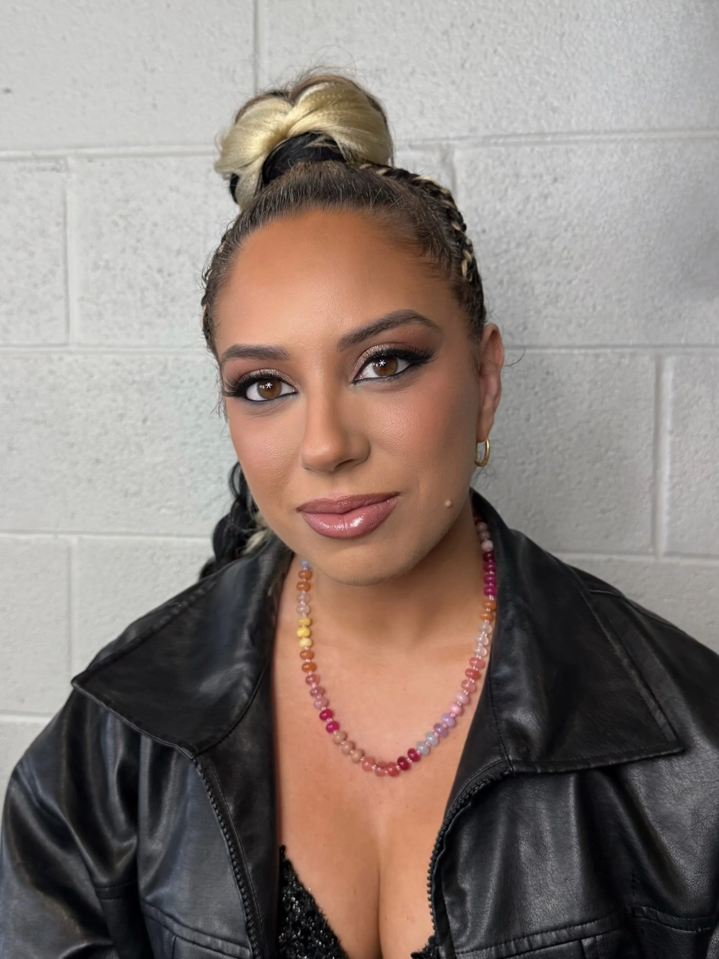 Close-up of a woman with styled hair, makeup, and jewelry, wearing a black leather jacket, against a white brick wall.