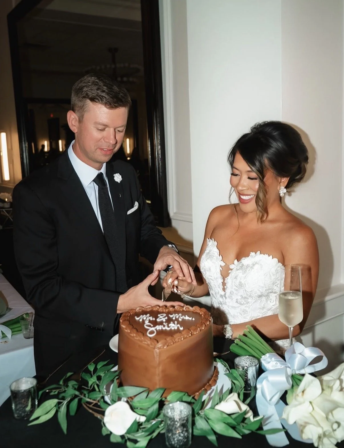 A bride and groom cutting a wedding cake together at their reception, with a decorated table featuring greenery and flowers, and a glass of champagne nearby.