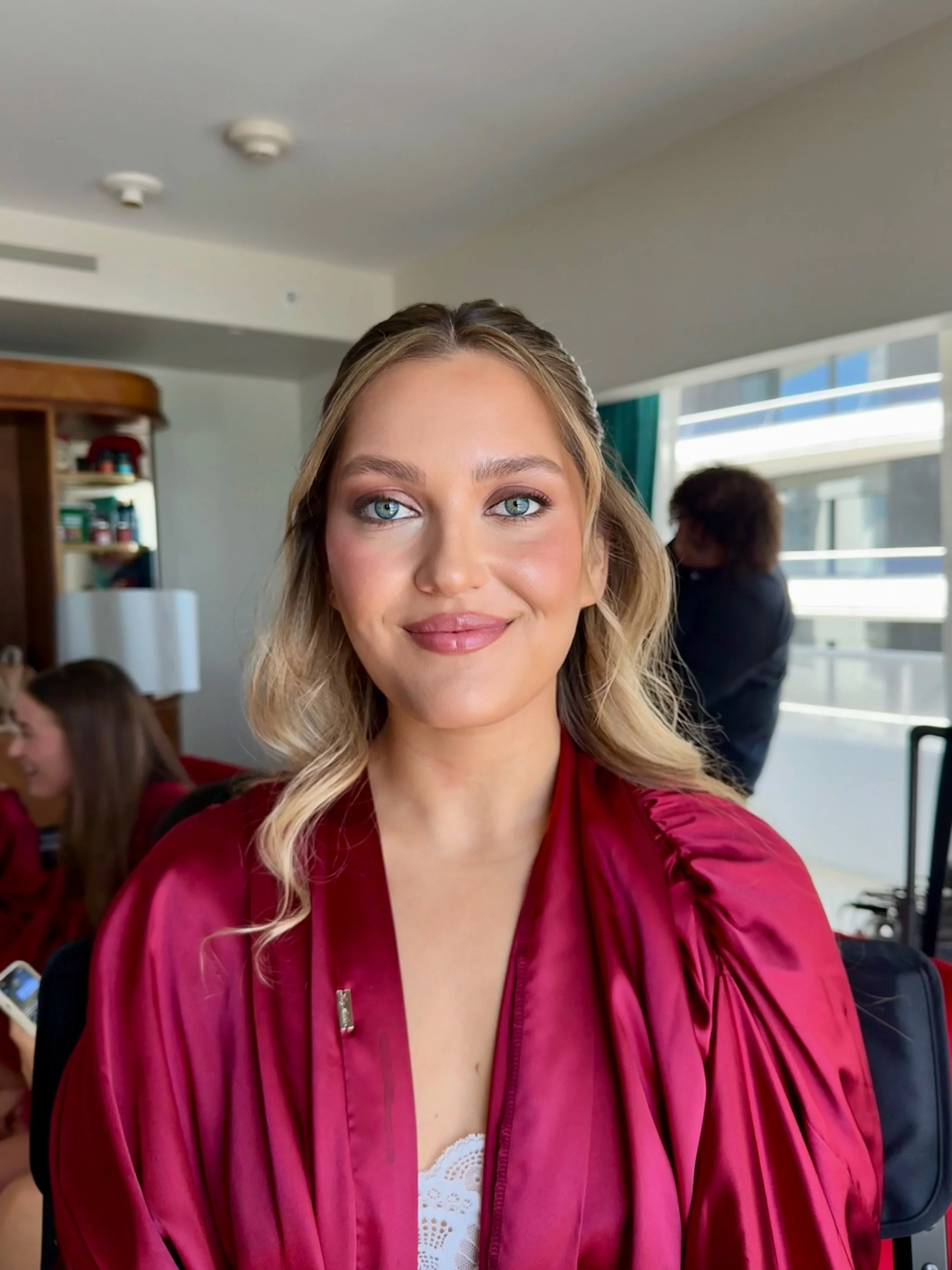 Young woman with blonde hair and blue eyes smiling, wearing a burgundy graduation gown, standing indoors with a bright window behind her.