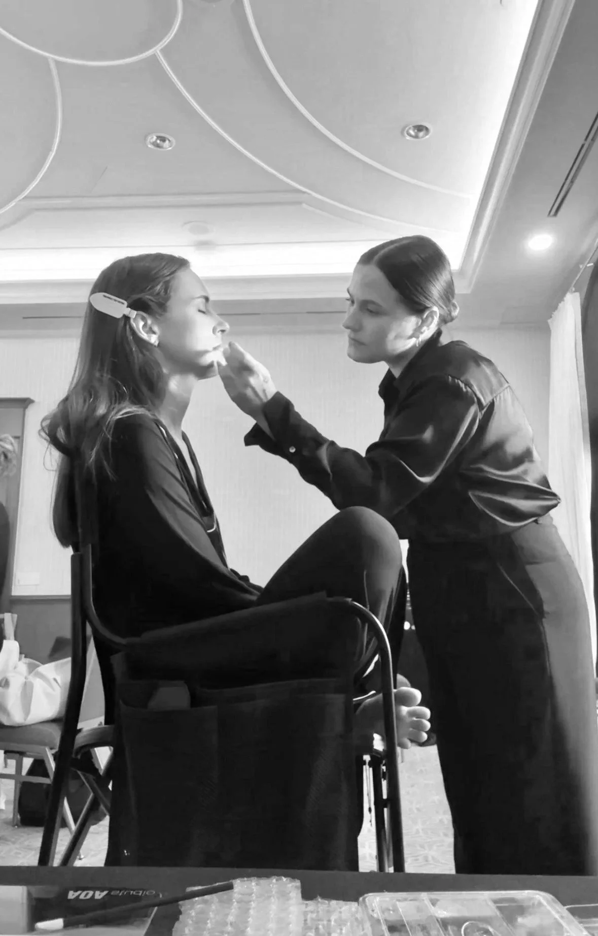 Woman sitting with eyes closed and woman standing applying makeup or face treatment in a well-lit room.