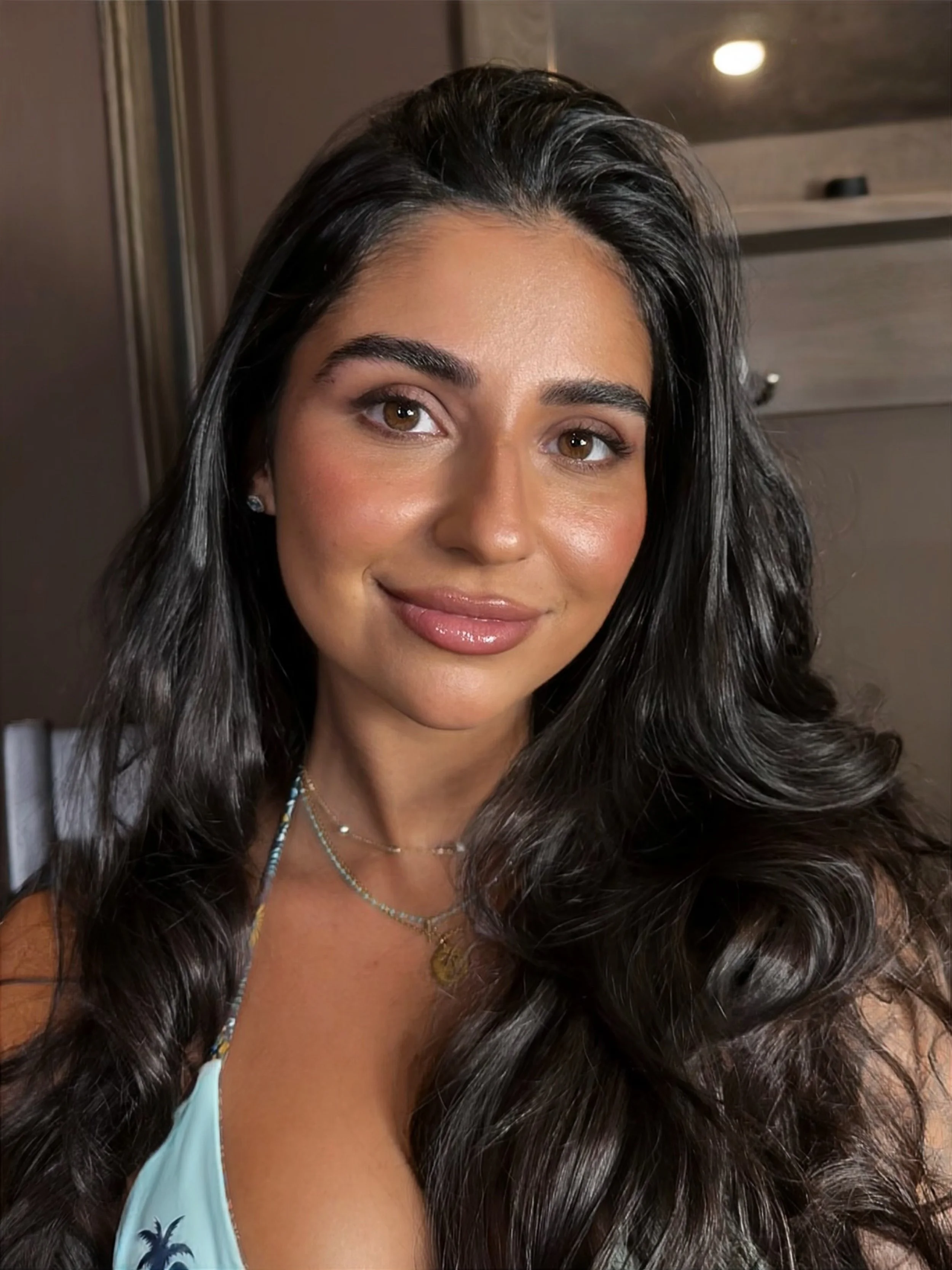 Close-up of a smiling woman with long dark wavy hair, wearing jewelry, with a blurred indoor background.
