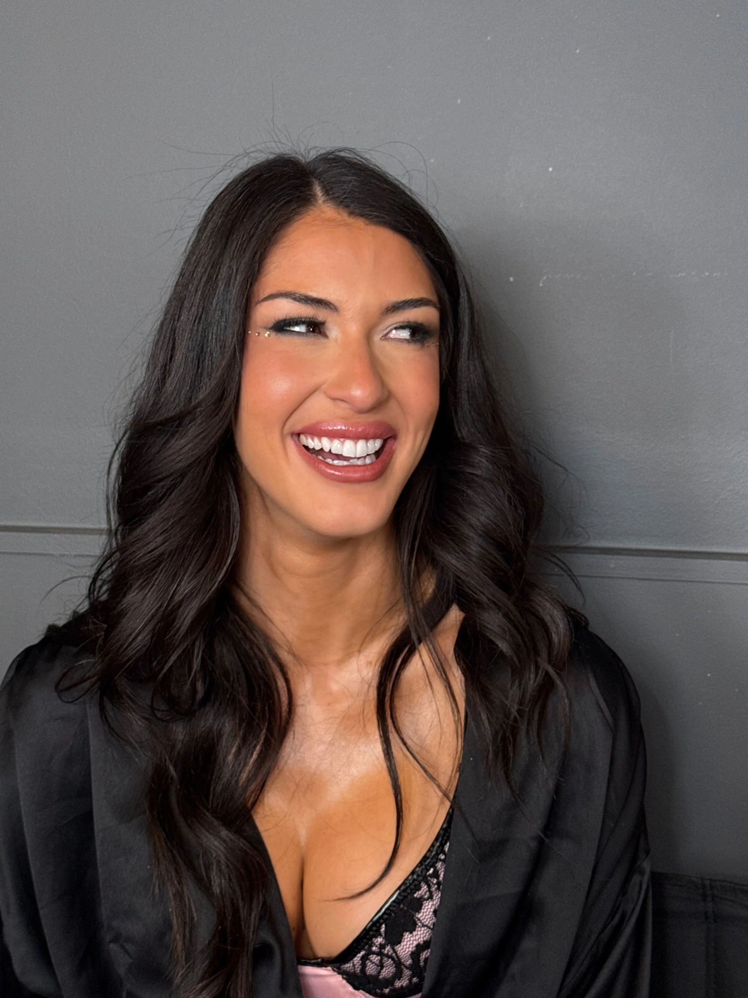 A woman with long dark hair, smiling and showing her teeth, wearing makeup, a black top with lace detail, and a bra strap visible. She stands against a gray wall.