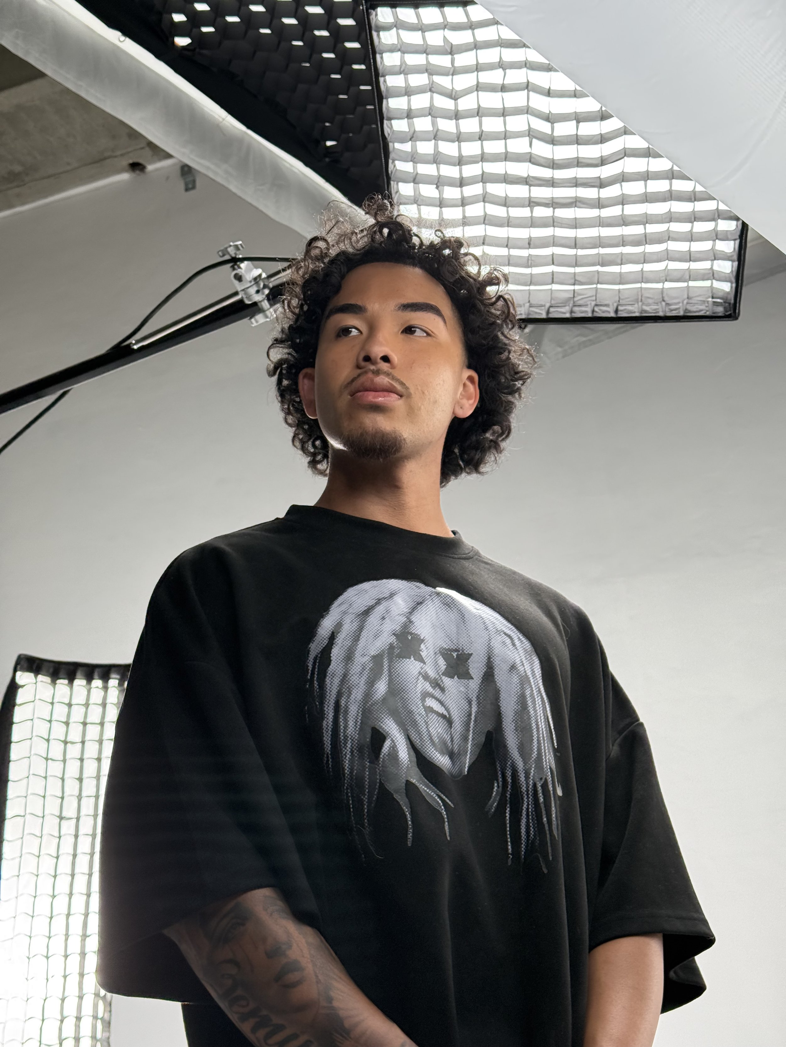 A young man with curly hair and tattoos on his arm stands in a photography studio, looking slightly away from the camera, with large studio lights and black soundproofing panels visible overhead.