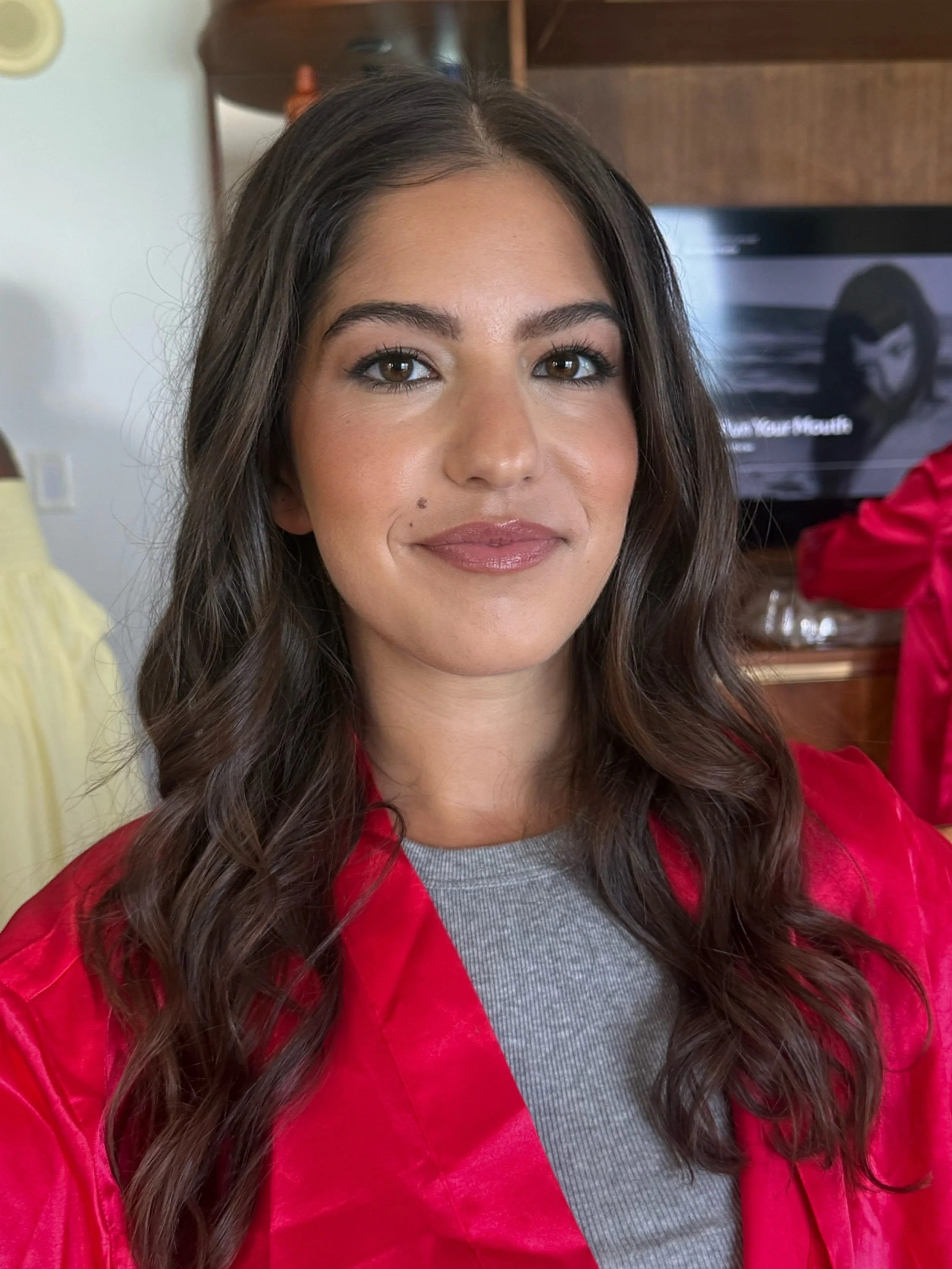Close-up of a woman with long, wavy brown hair, wearing makeup, a red jacket, and a gray top, smiling softly in a room with a TV and wooden furniture in the background.