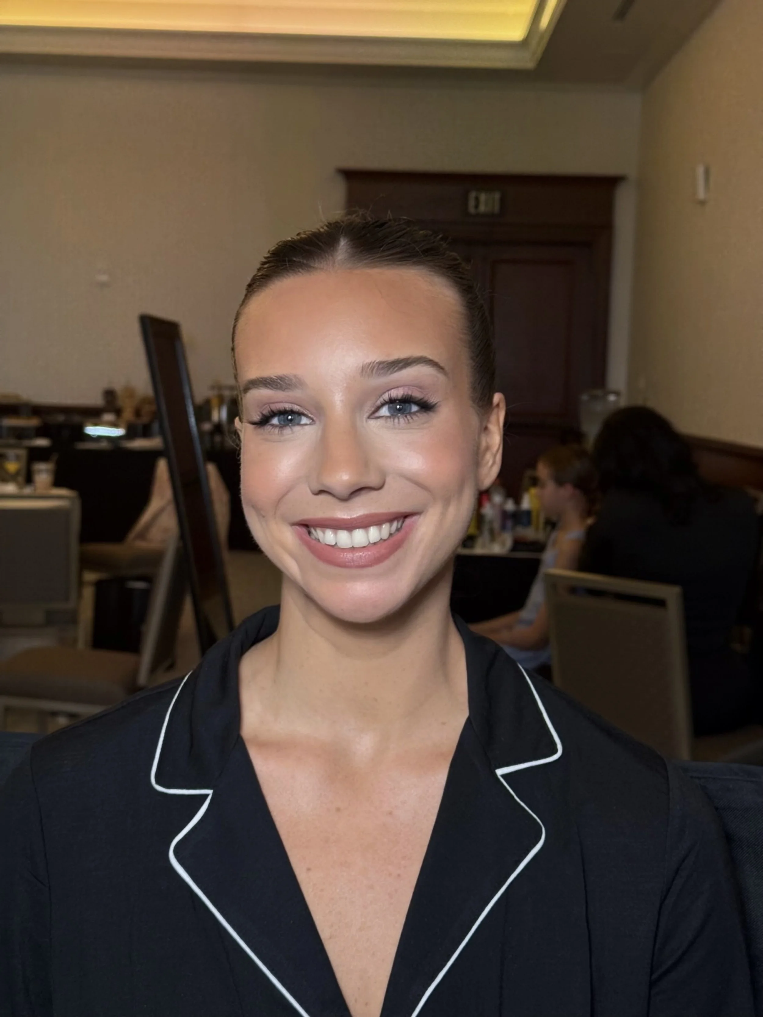 Close-up of a woman with blonde hair styled in a bun, smiling, wearing black pajamas with white piping, in a room with beige walls and wooden accents, and people in the background.