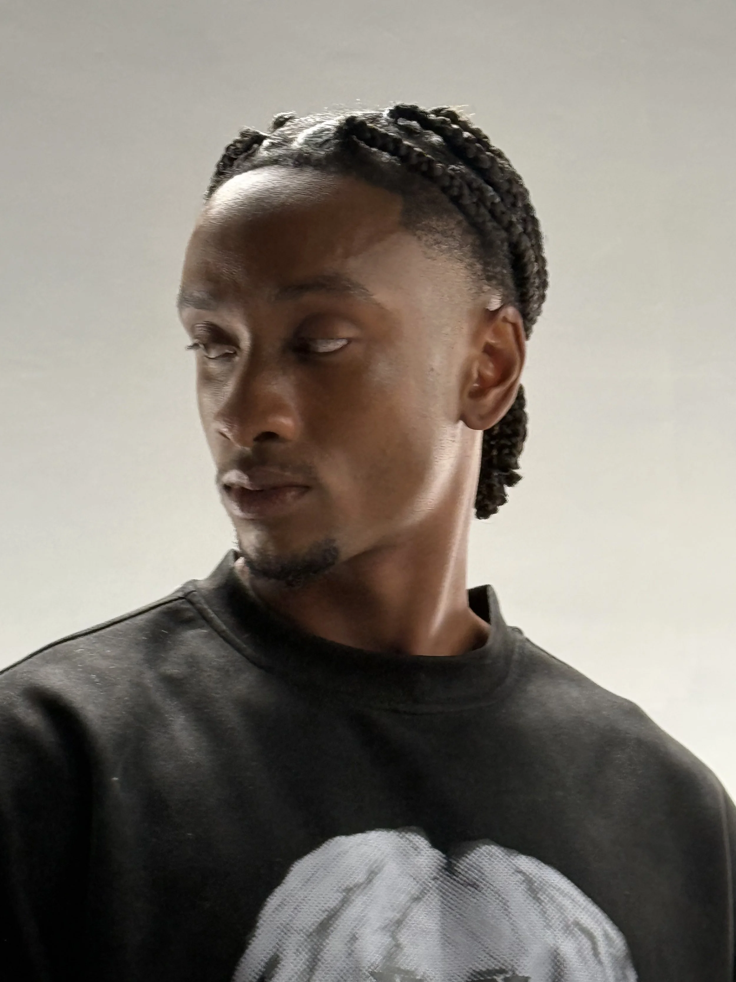 A person with dark skin, styled with braids, is looking downward and to the side, wearing a black sweatshirt, against a plain light-colored wall.