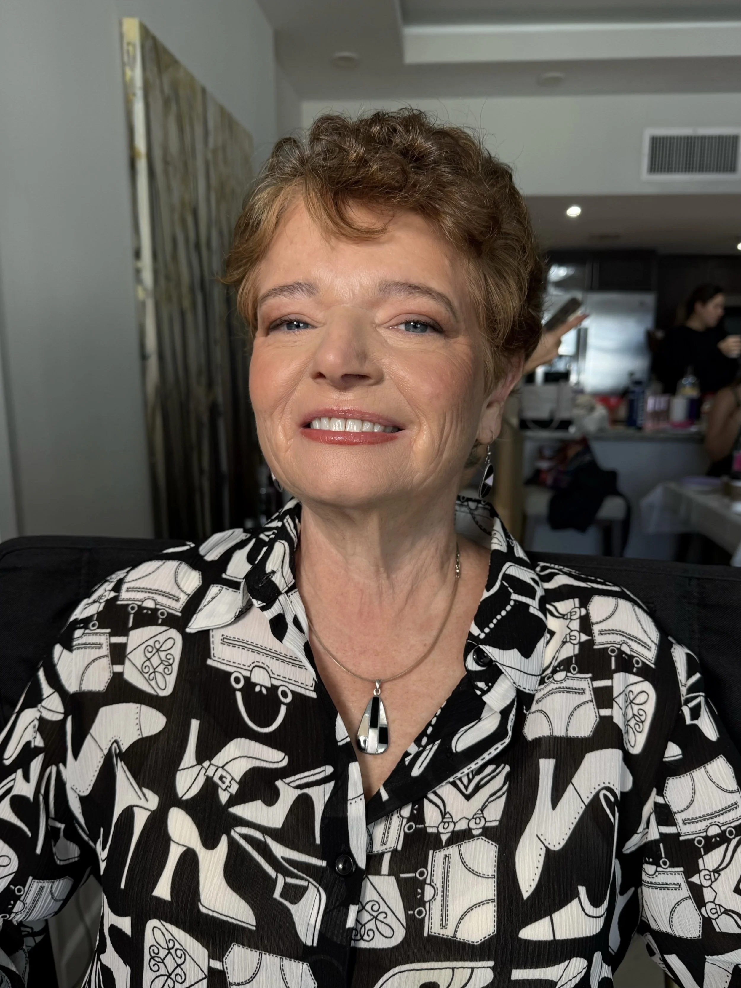 Close-up of a smiling woman with short, curly brown hair and blue eyes, wearing a black and white patterned shirt, sitting indoors with a blurred background of people and modern interior design.