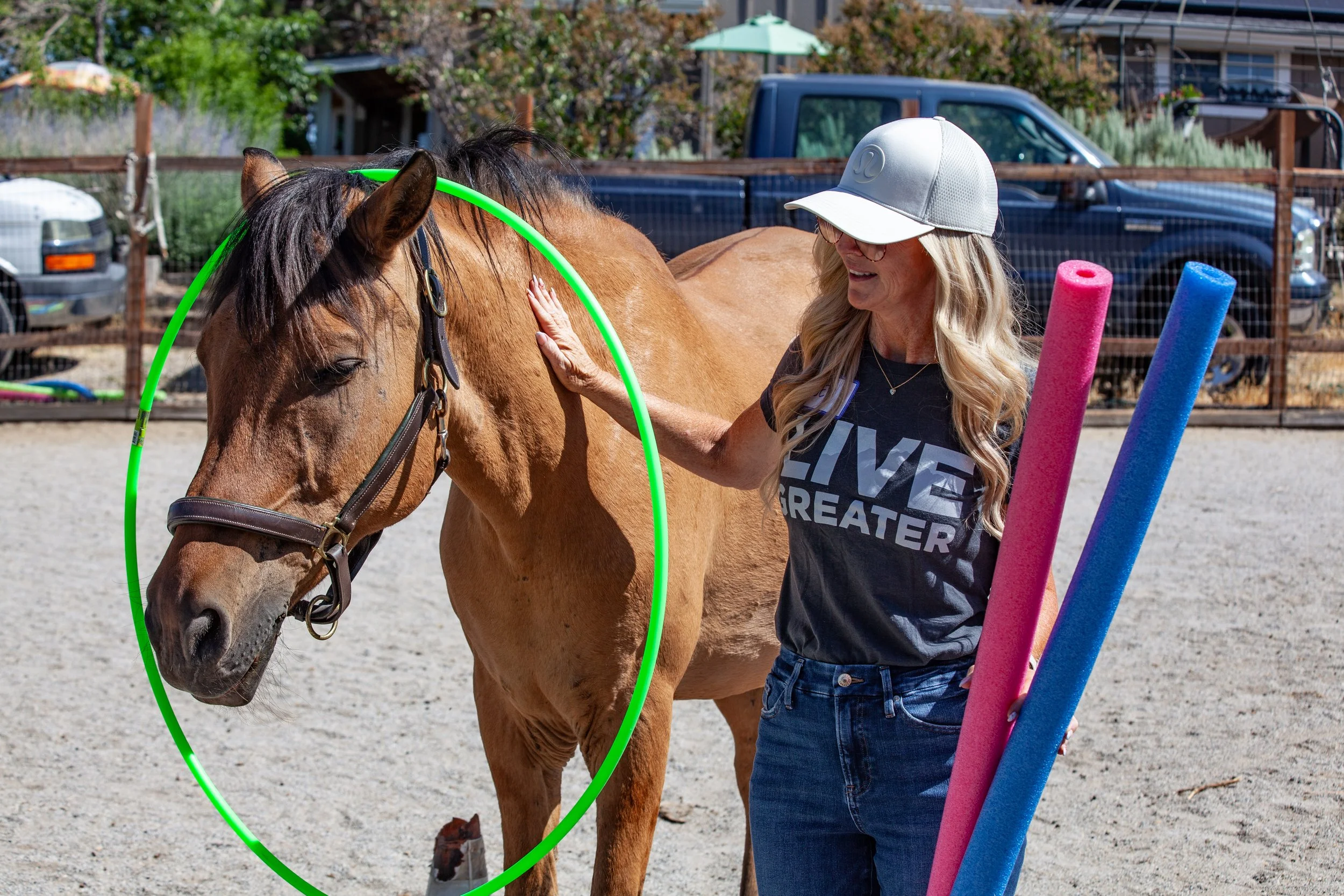 Woman standing next to horse wearing a green hula hoop holding two pool noodles and petting the horse's neck.