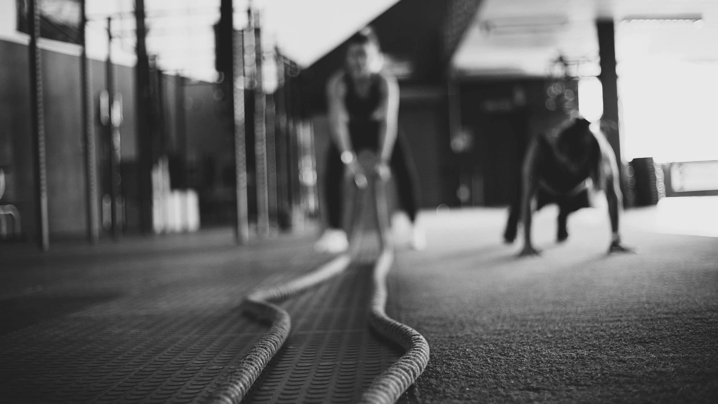 Two people exercising with battle ropes in a gym, black and white photo, one person blurred in the background and one in a push-up position.