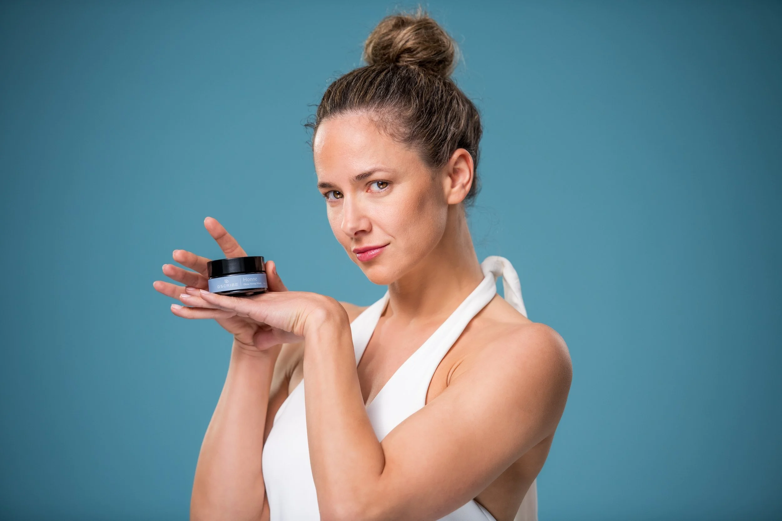 Person holding a small black jar of skincare cream against a blue background, wearing a white top.