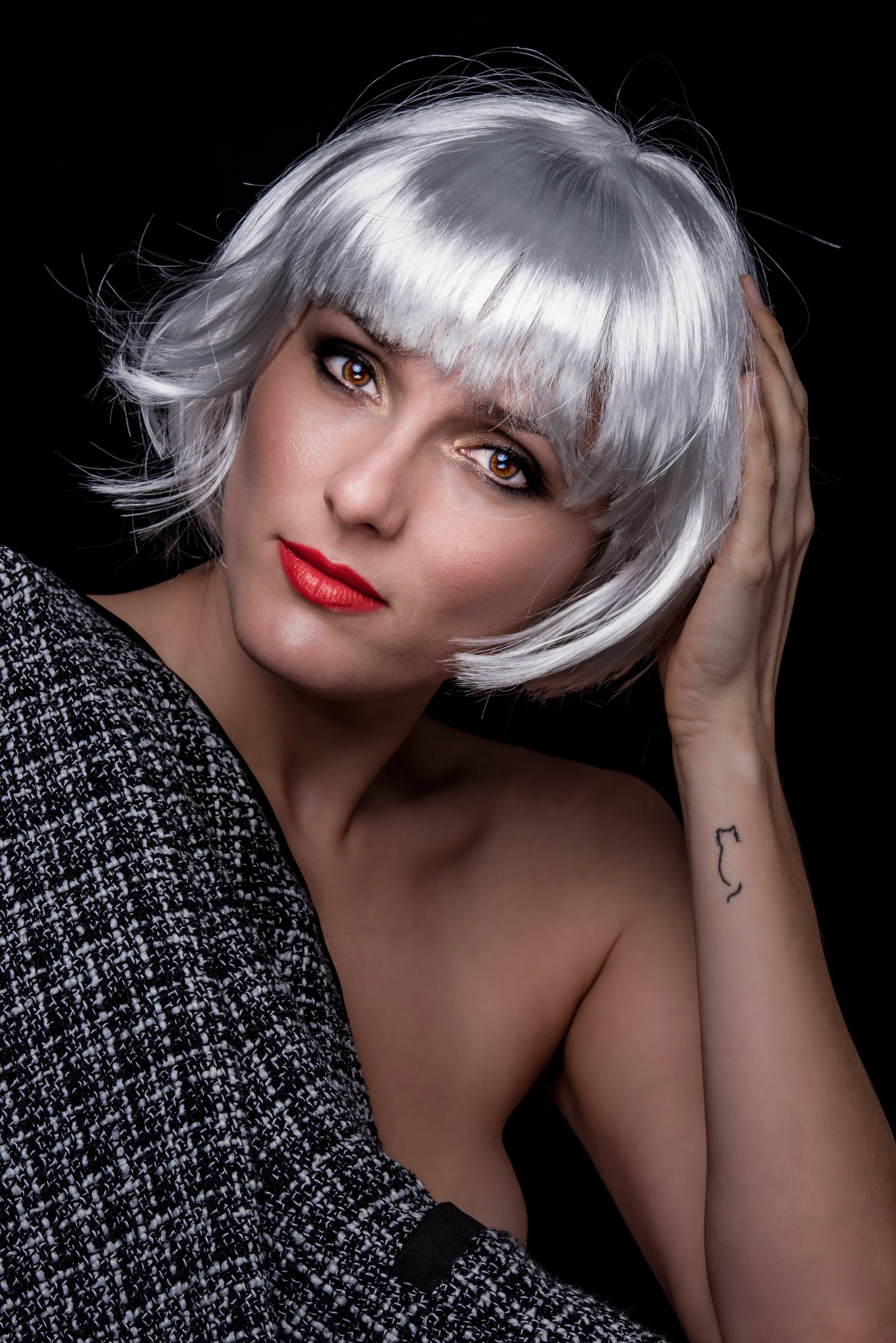 Woman with short silver hair and red lipstick wearing a black and white textured fabric over her shoulder, posing against a black background.