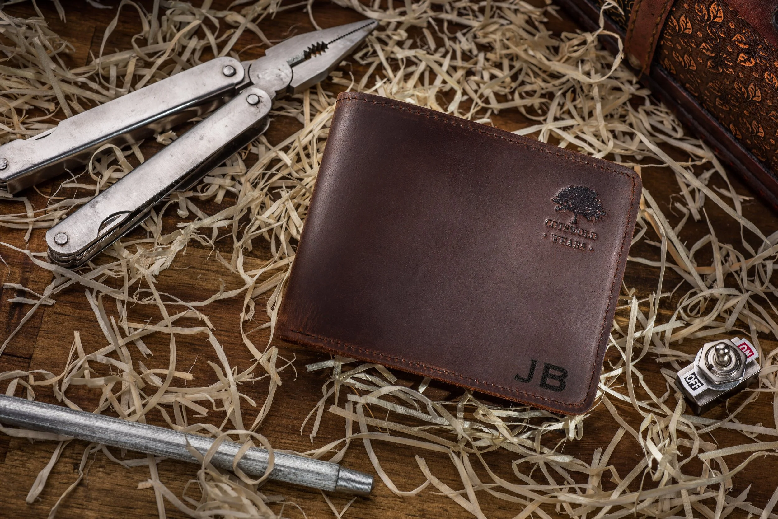 Brown leather wallet on wooden surface, surrounded by straw, with a multitool, metal rod, and a toggle switch nearby.