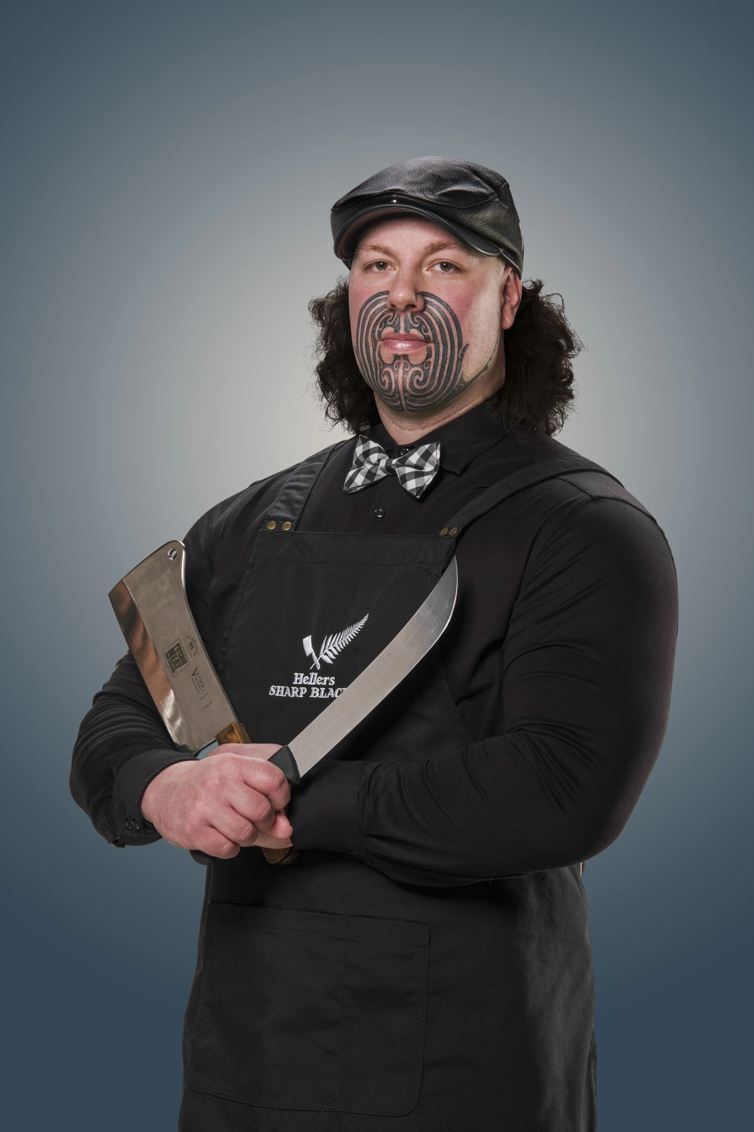 Person wearing a black apron and cap, holding two large knives, with facial tattoos.