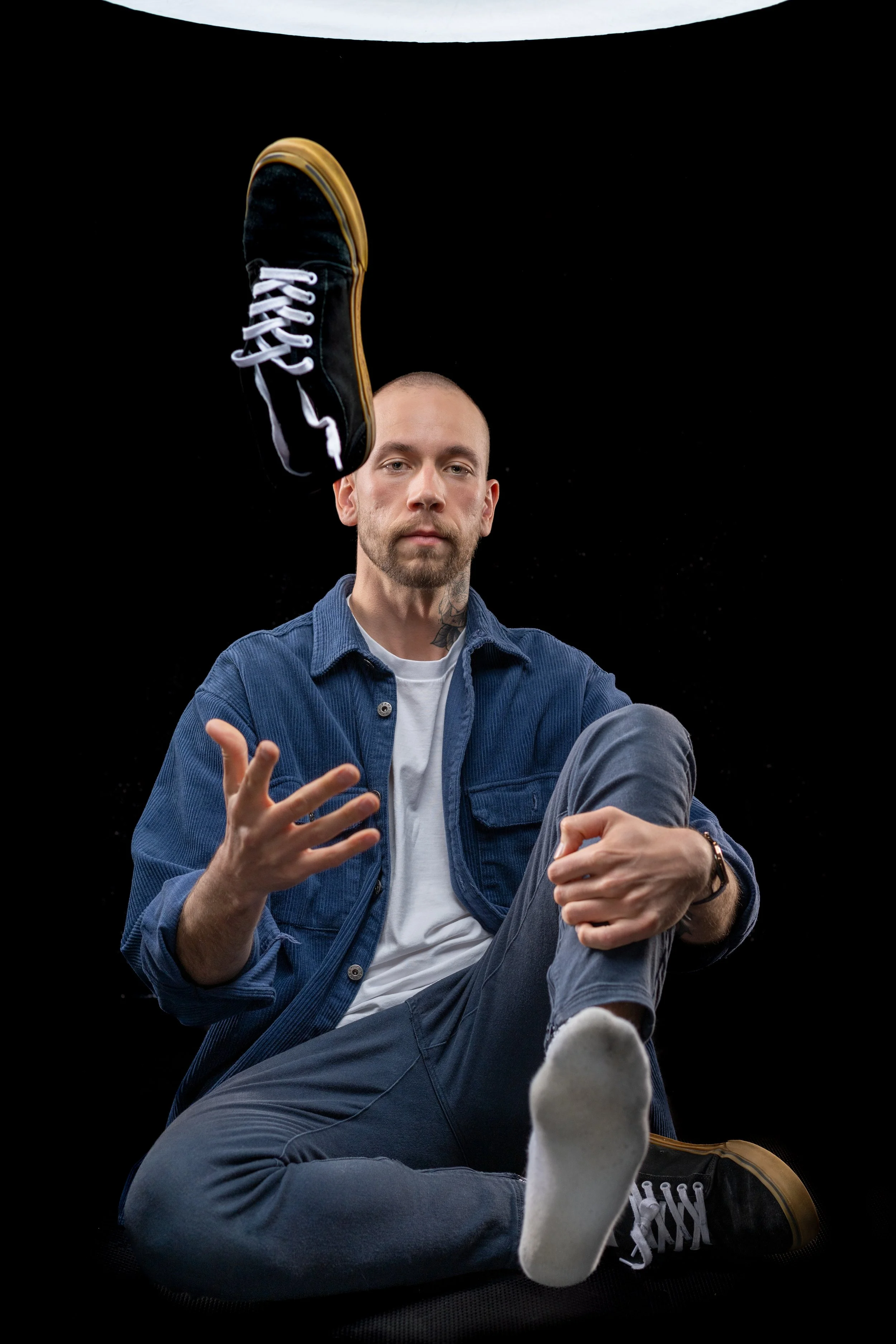 Man sitting, tossing a black sneaker, wearing denim outfit, barefoot, against black background.