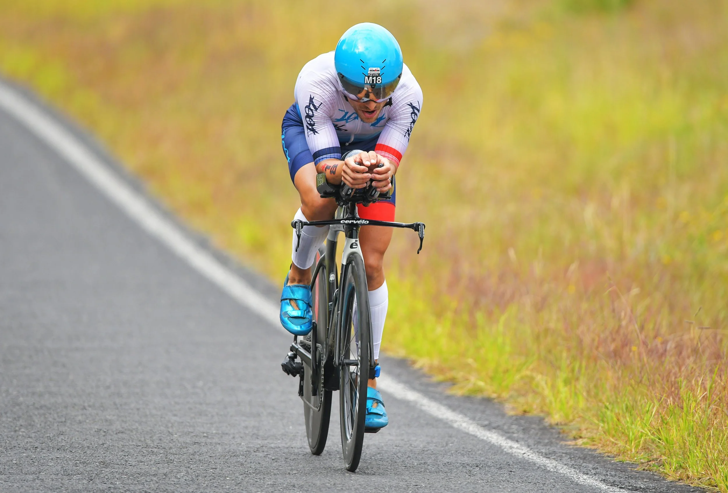 Cyclist in blue helmet and white cycling suit riding a time trial bike on a rural road.