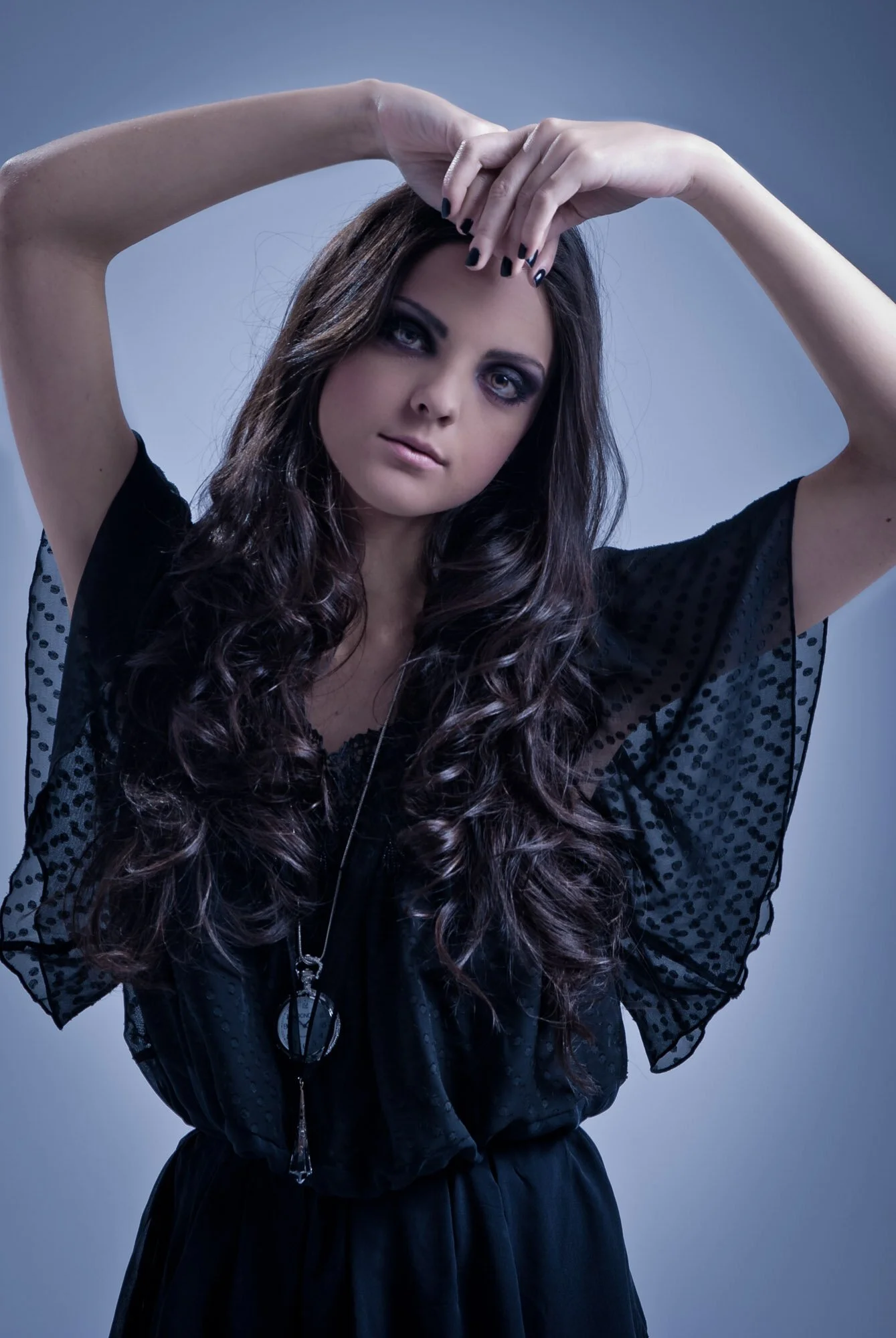 Woman in black dress with dark makeup, long curly hair, posing with arms raised against a blue background.
