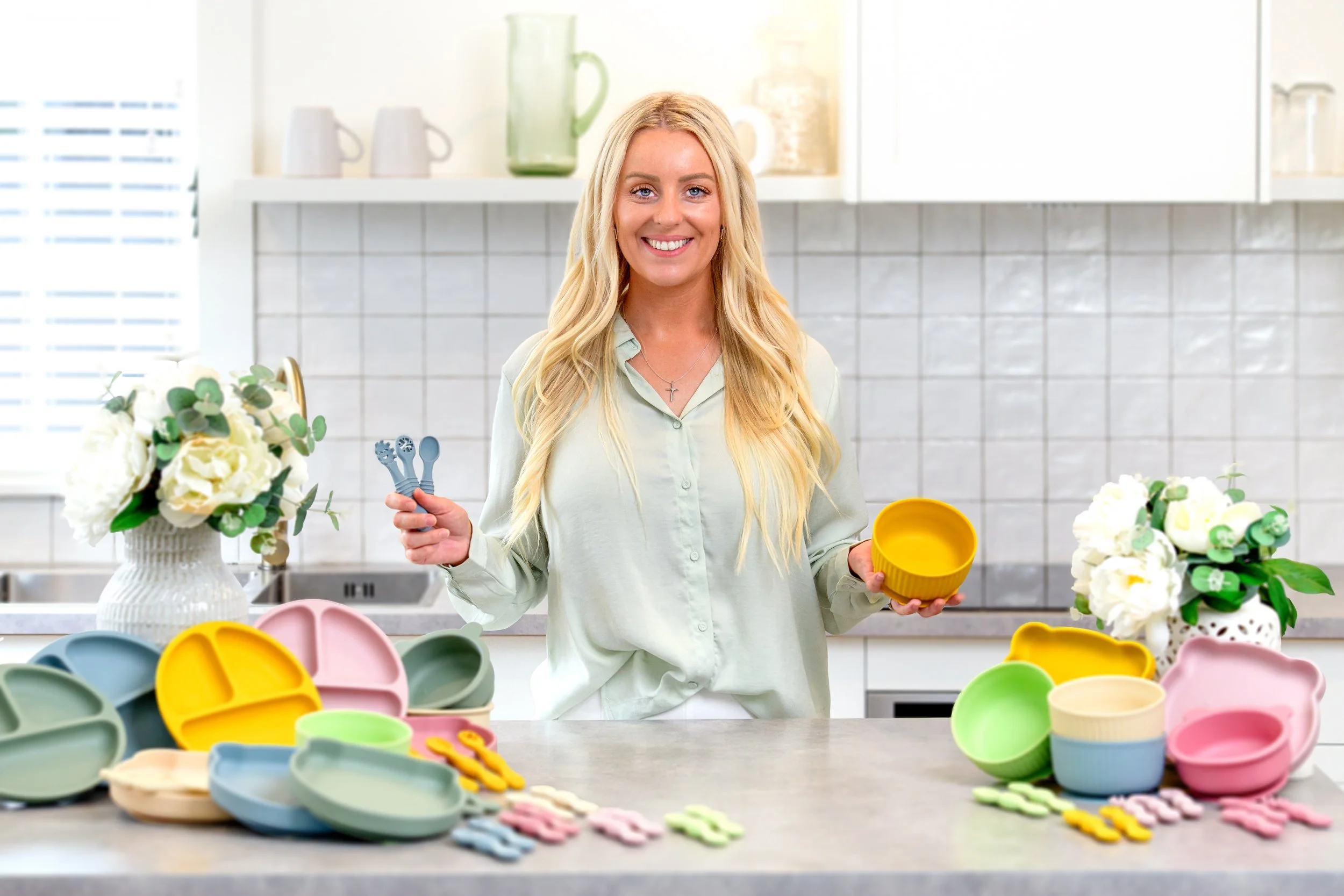 A person in a kitchen holding children's utensils and a yellow bowl, with various colorful children's dishes and utensils spread out on the countertop.