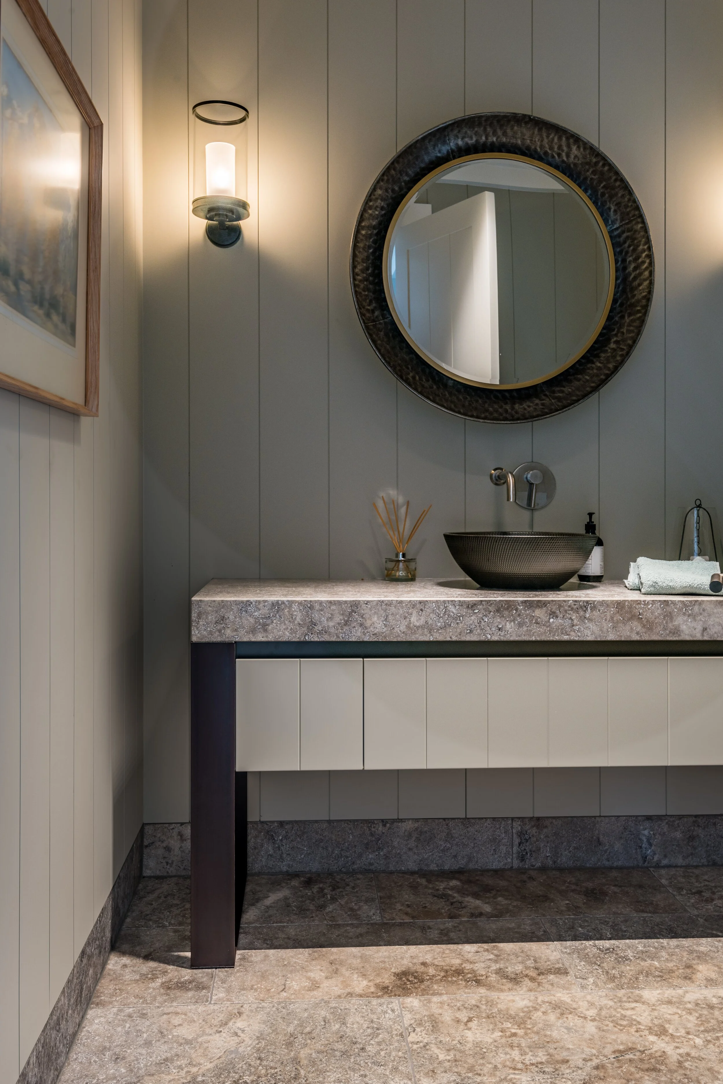 Modern bathroom vanity with round mirror, stone countertop, vessel sink, wall sconce, and framed wall art.
