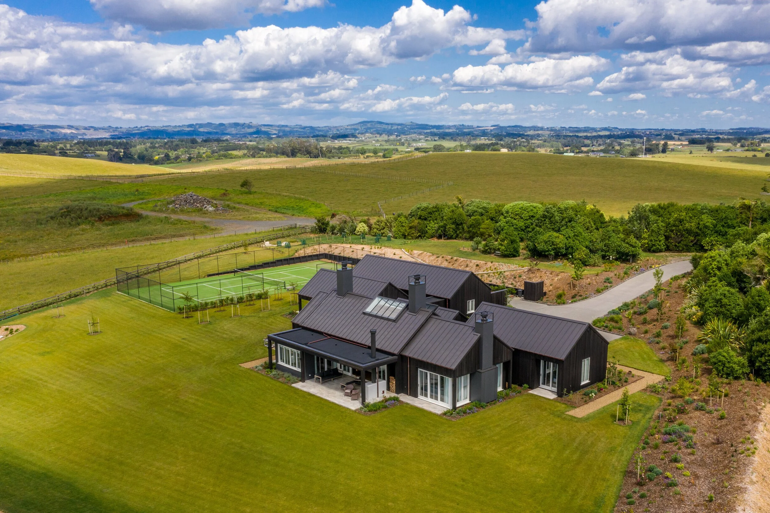 Aerial view of a modern house with a black roof and surrounding manicured lawn, adjacent to a fenced tennis court, set in a rural landscape with fields and distant hills under a cloudy sky.