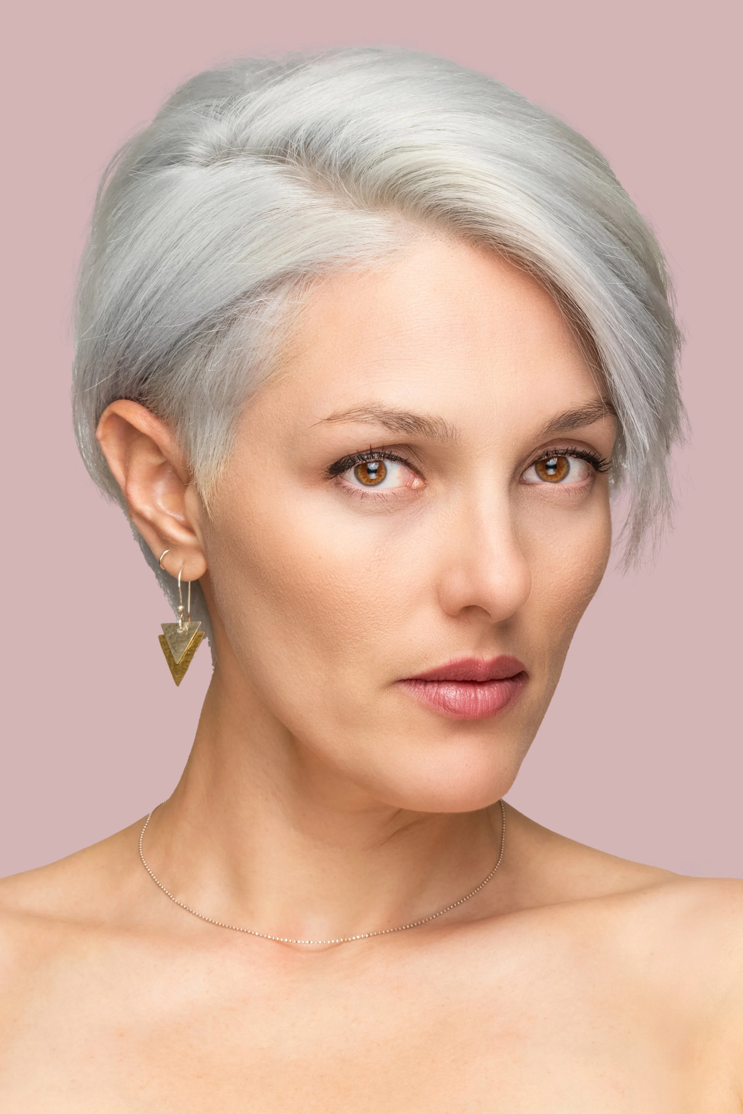 Woman with short gray hair, wearing triangle earrings and a delicate necklace, against a pink background.