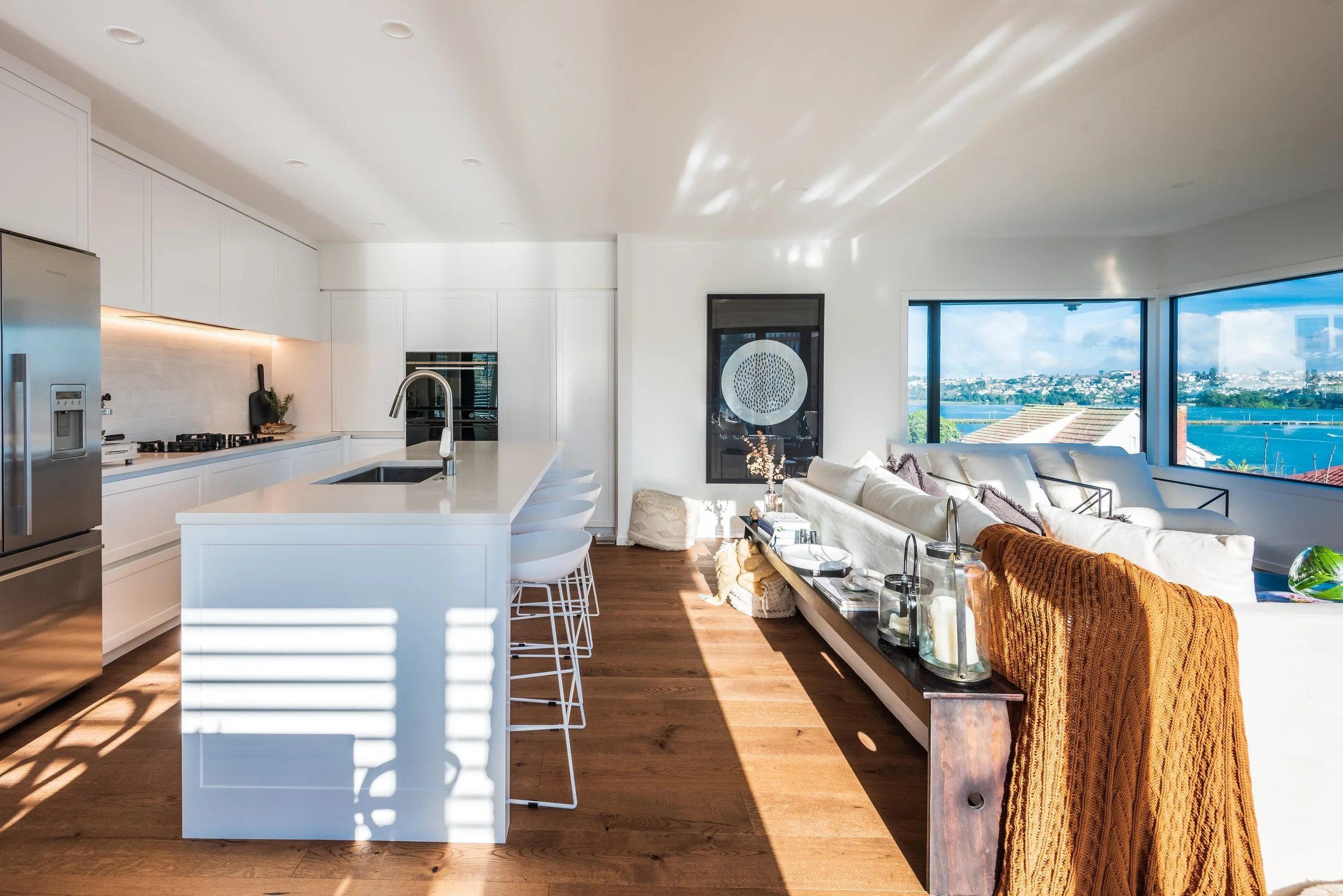 Modern kitchen and living area with white cabinets and island, hardwood floors, large windows, and ocean view.