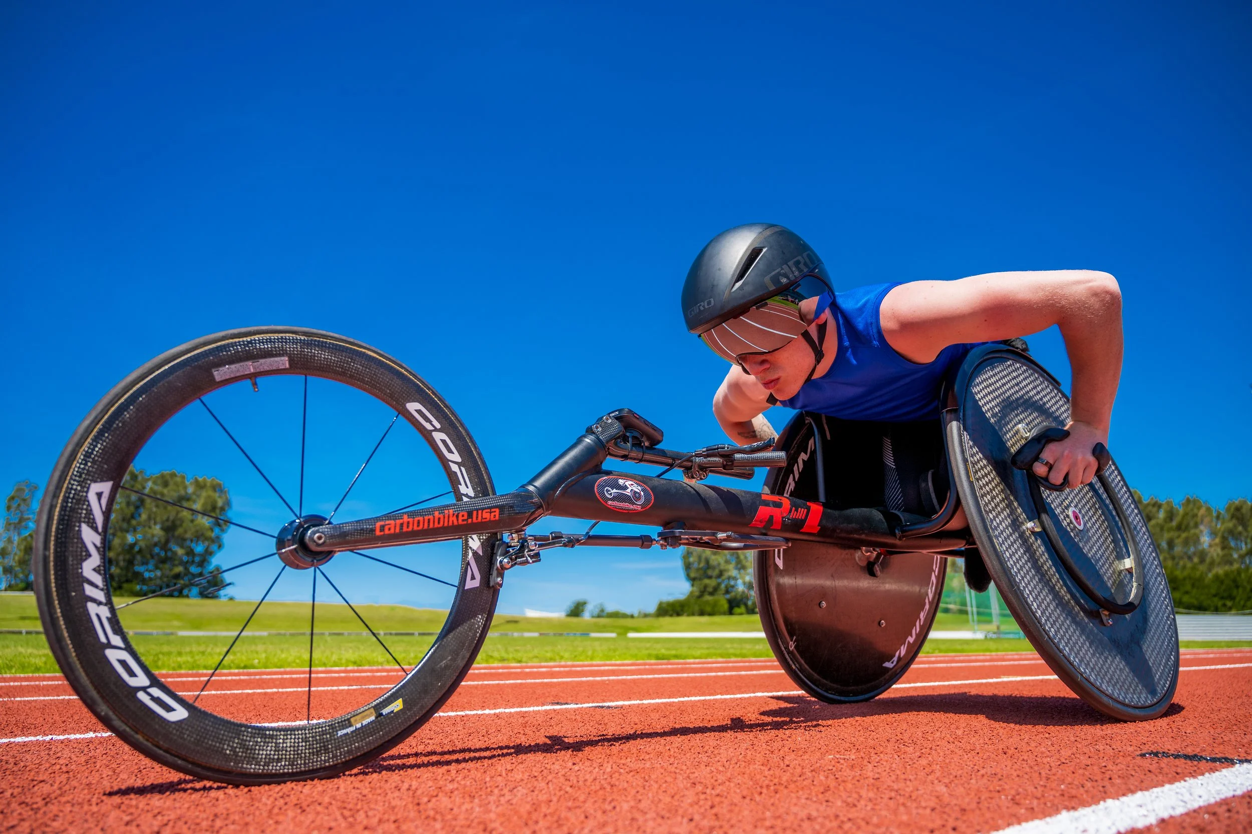 Athlete in a racing wheelchair on a track, wearing a helmet and sunglasses, under a clear blue sky.