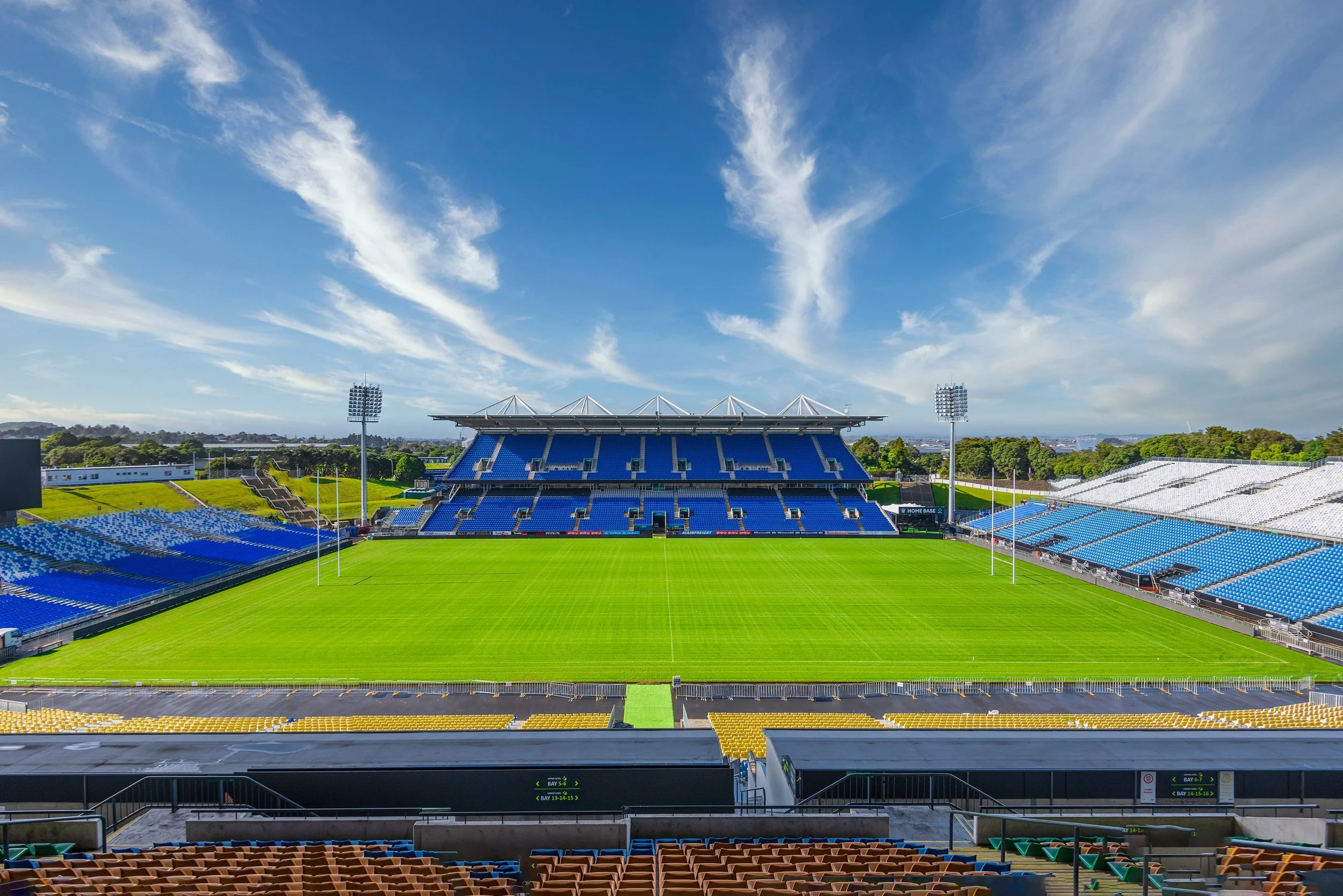 Rugby stadium with blue and white seating, green field, goal posts, and clear sky.