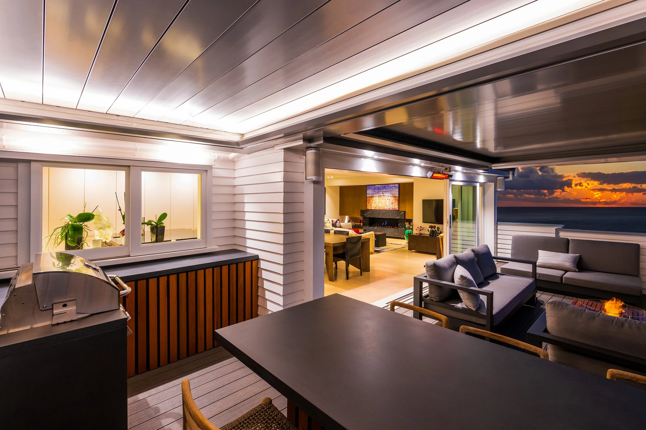 Modern outdoor patio with a grill, seating area, and view of a sunset over the ocean in the background.