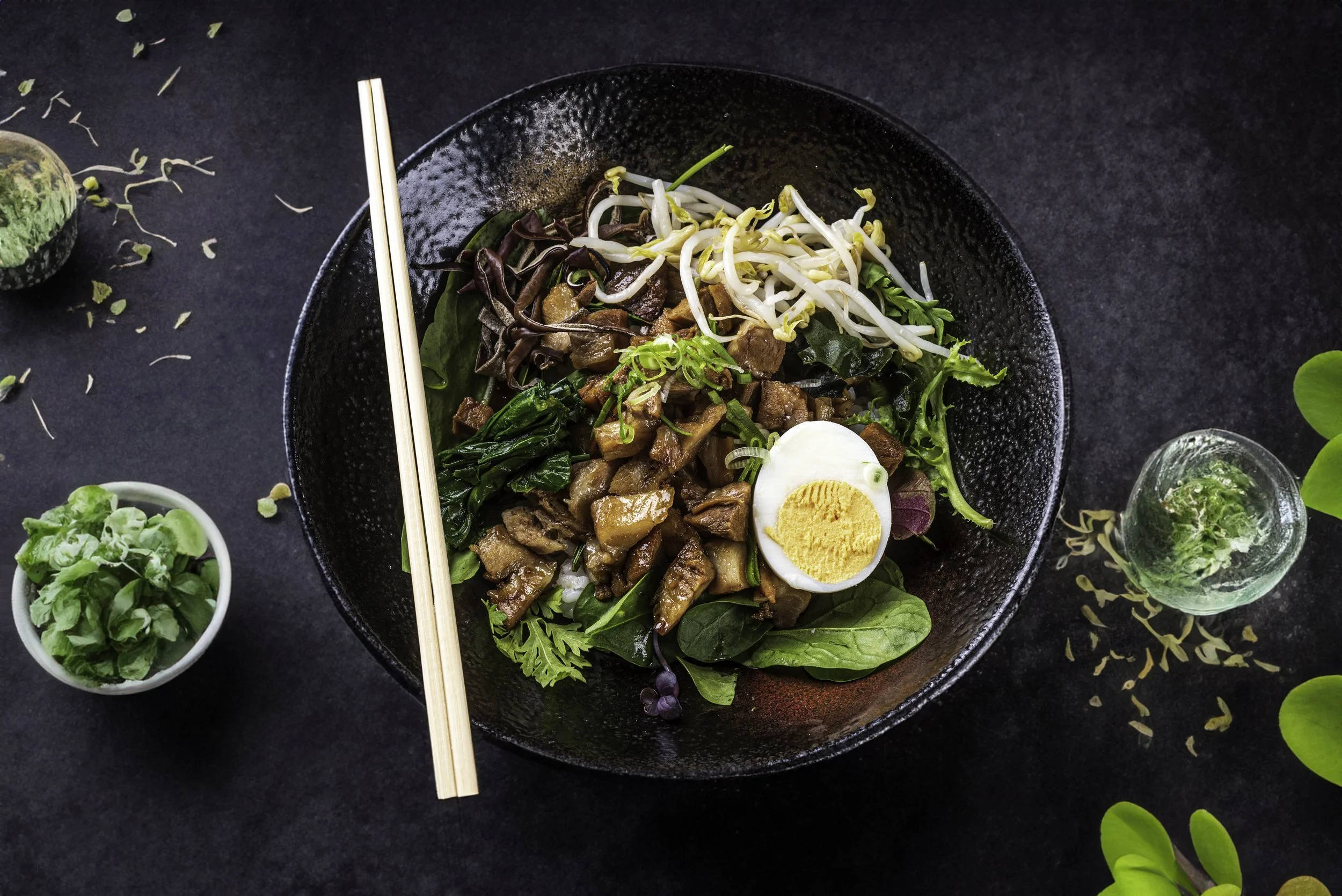 Bowl of mixed vegetables, sautéed mushrooms, boiled egg, and sprouts with chopsticks on a dark background, accompanied by a small bowl of fresh greens and scattered microgreens.