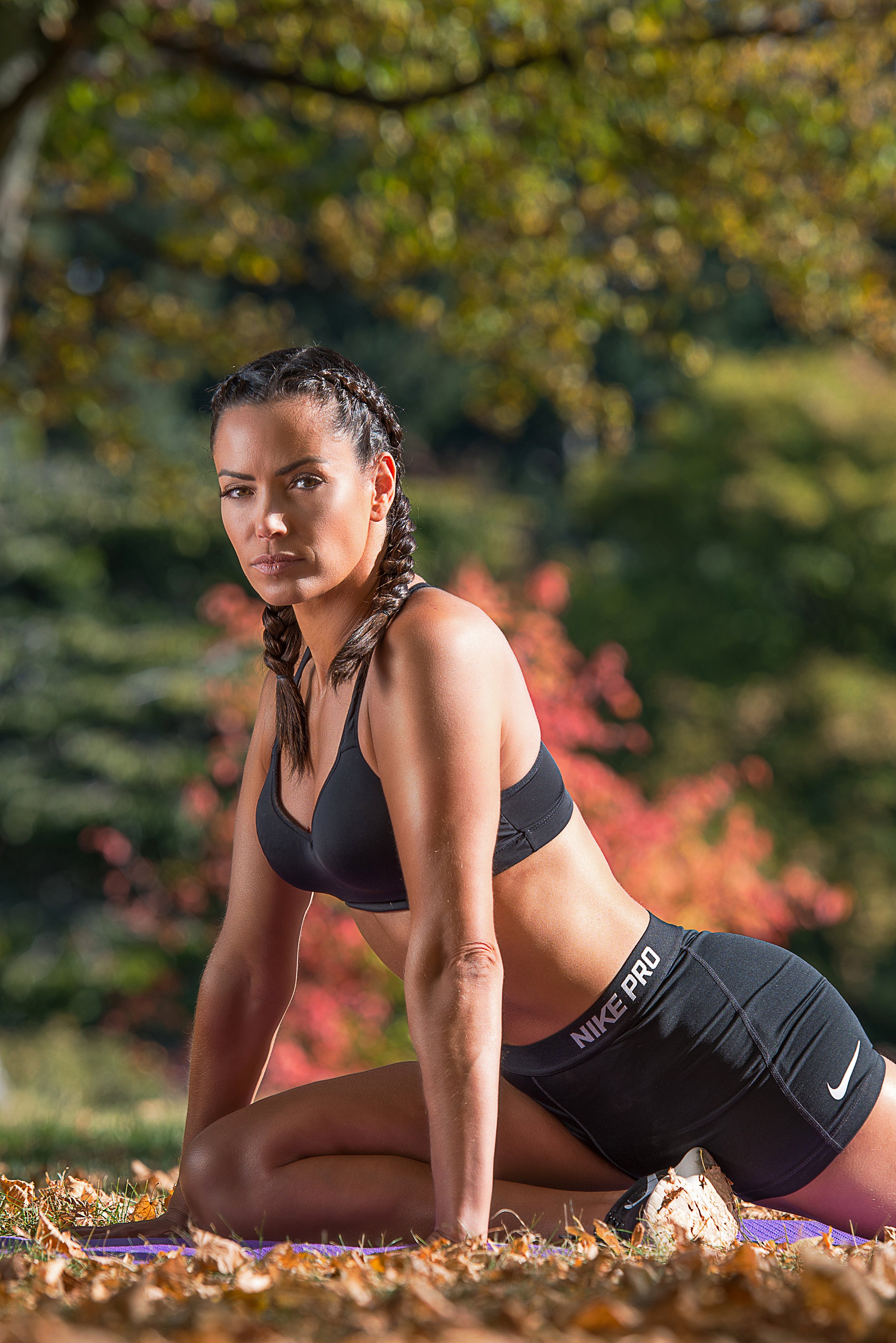 A woman in workout attire, with braided hair, performing a yoga pose outdoors in a park with autumn leaves.