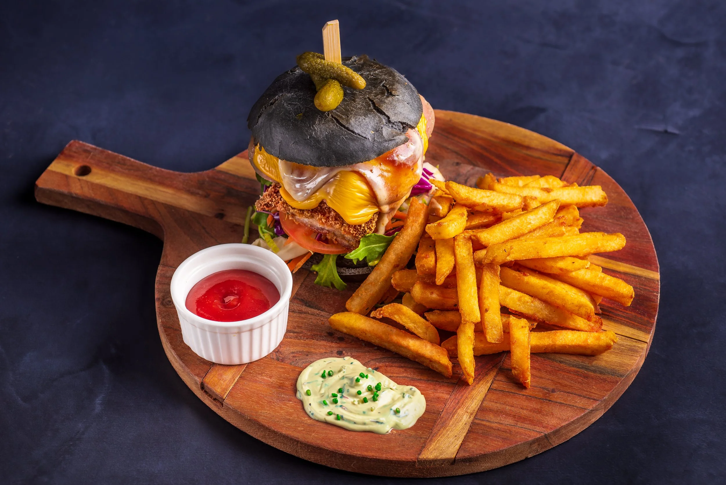 Gourmet charcoal bun burger with cheese and toppings on a wooden serving board, accompanied by golden fries and sauces, including ketchup and mayonnaise with herbs.