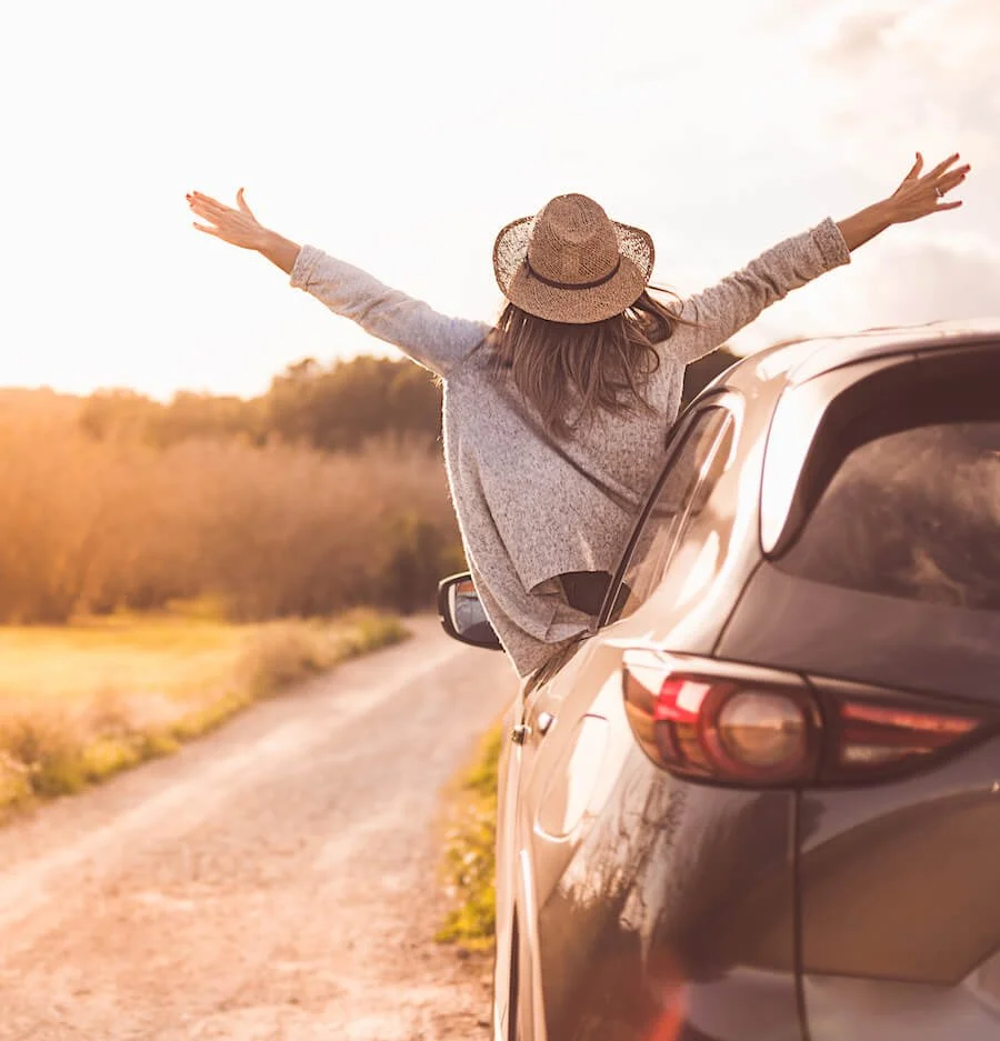 happy woman parked on a country road with her head and arms outstretched out the window of the car
