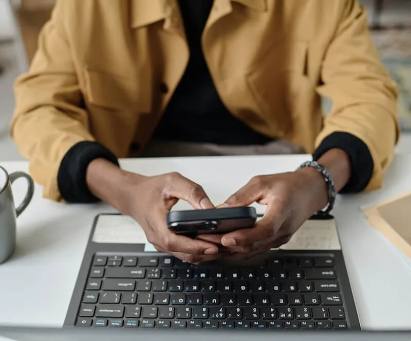 man's hands holding smart phone next to laptop computer