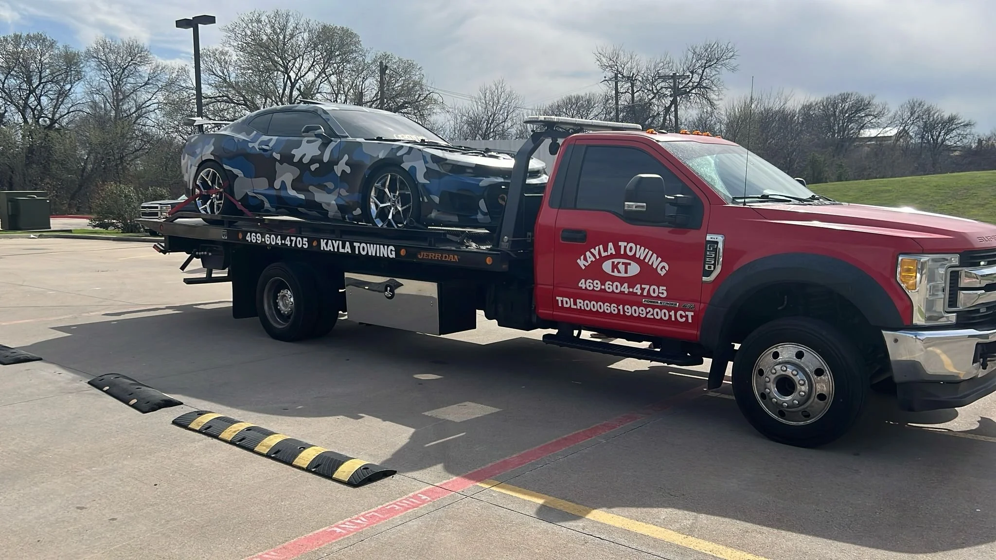 Red tow truck carrying a camouflage-patterned sports car in a parking lot on a cloudy day.