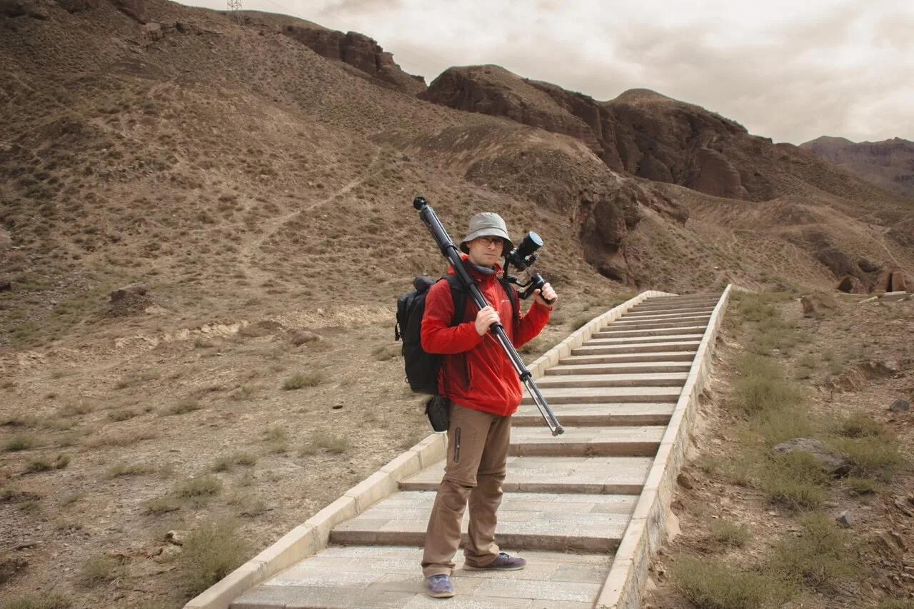 Person in outdoor gear holding a camera with telescope on a trail in a mountainous desert landscape under cloudy sky.