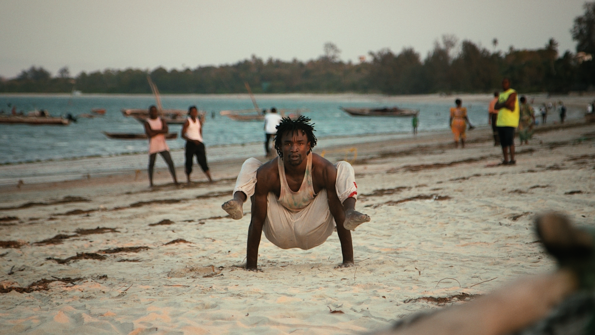 A black man on a busy beach with people and wooden boats in the background holds his body up with his hands while in a contorted position