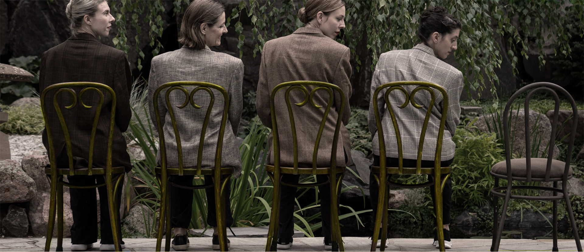 Four women sitting on chairs outdoors, facing away from the camera, with lush green plants and rocks in the background.