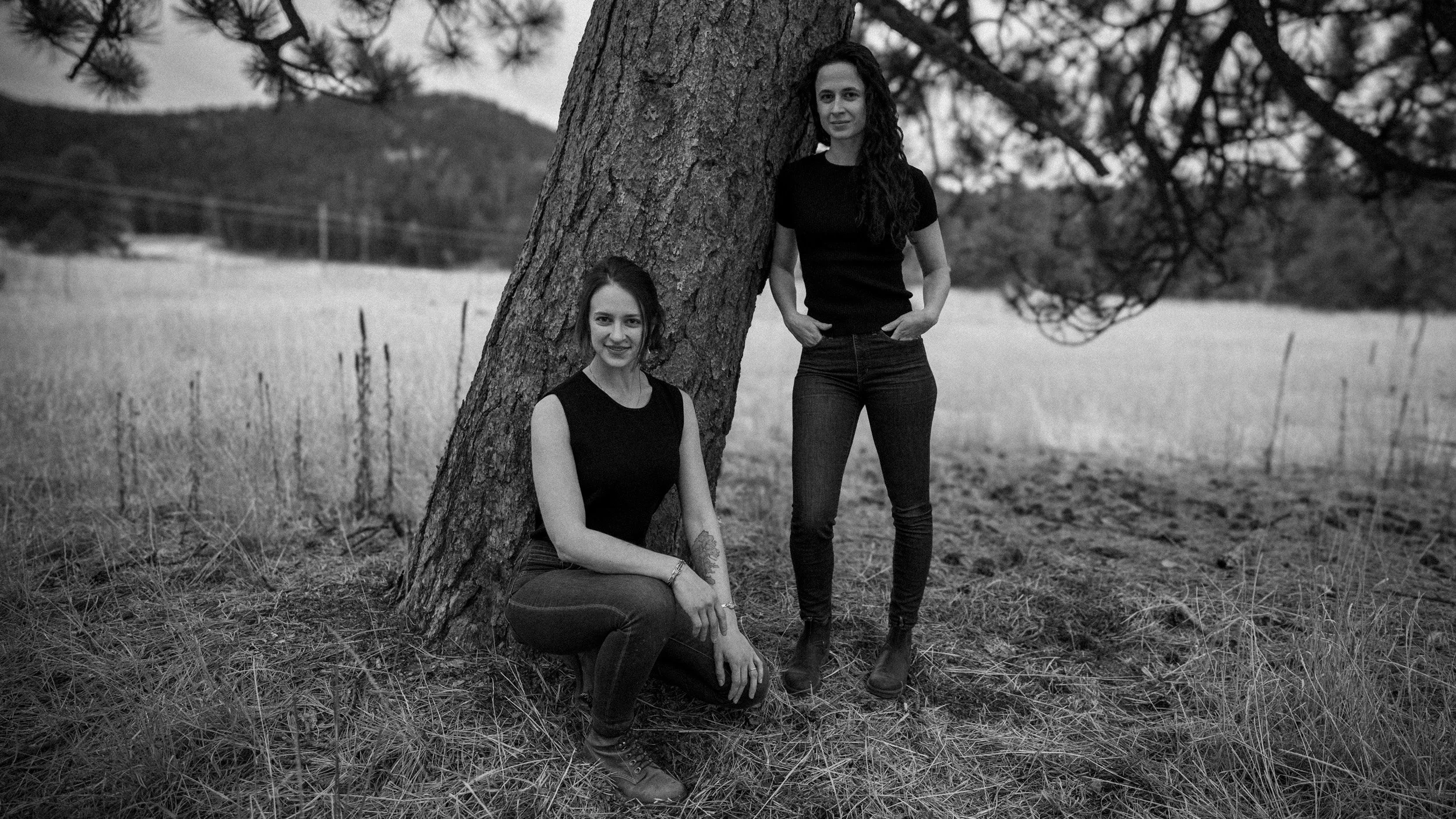 Two women with long curly hair, one sitting on the ground and the other standing, leaning against a large tree in an outdoor field.