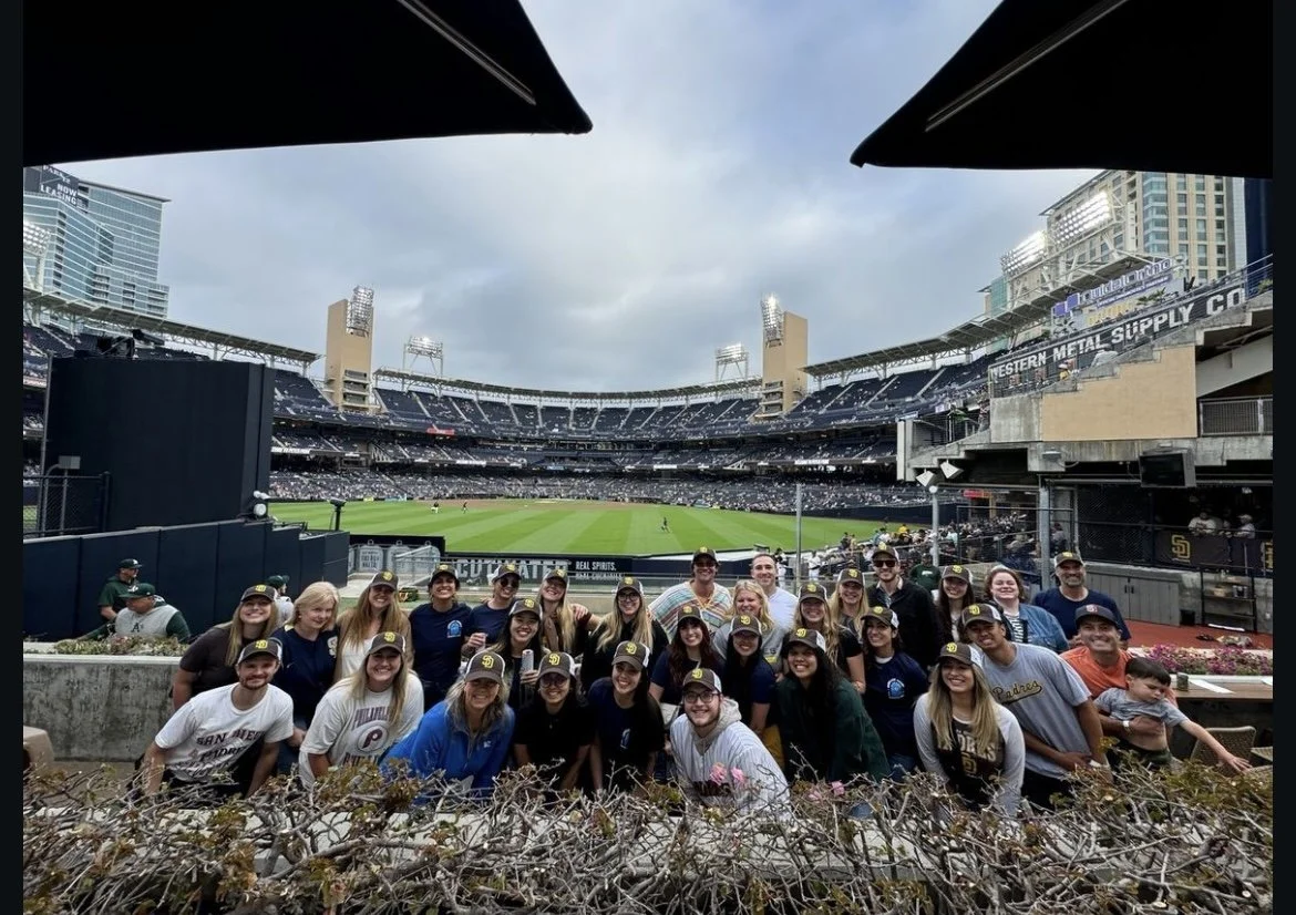 Group of people posing with a baseball stadium in the background during a game day.