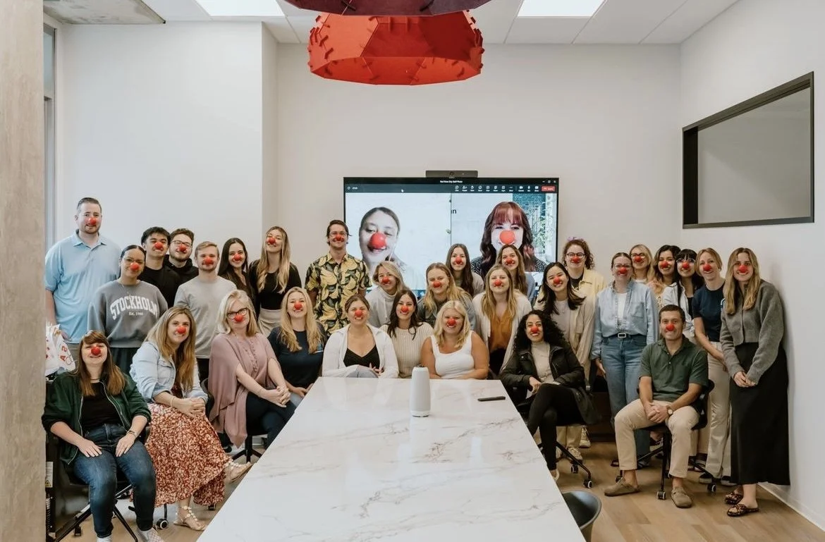 A group of people in an office setting, all wearing red clown noses, standing around a conference table. A screen in the background shows a video call with two participants also wearing red noses.