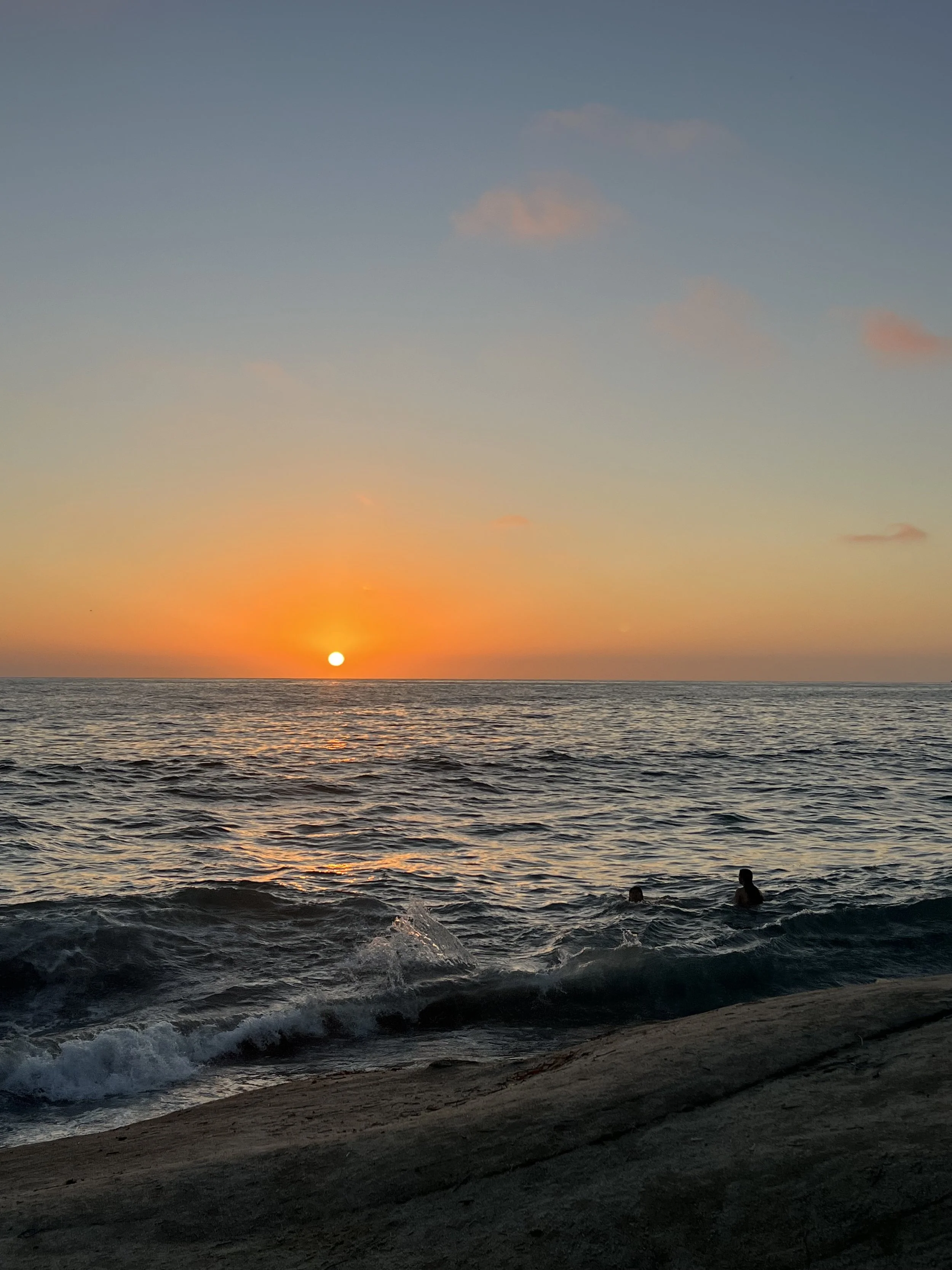 Sunset over ocean with two people swimming near rocky shore.