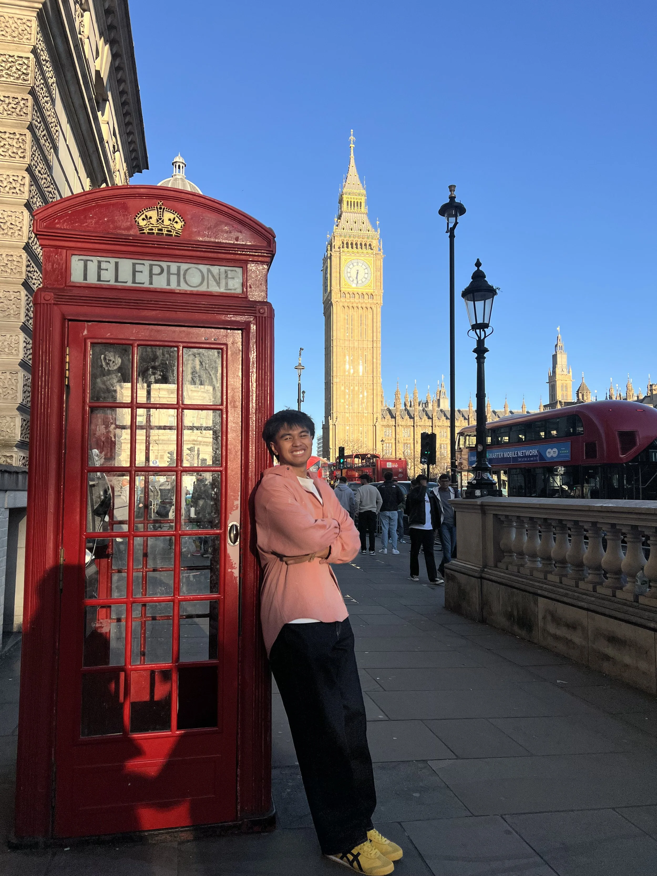 Person standing next to a red telephone booth with Big Ben clock tower in the background on a sunny day.