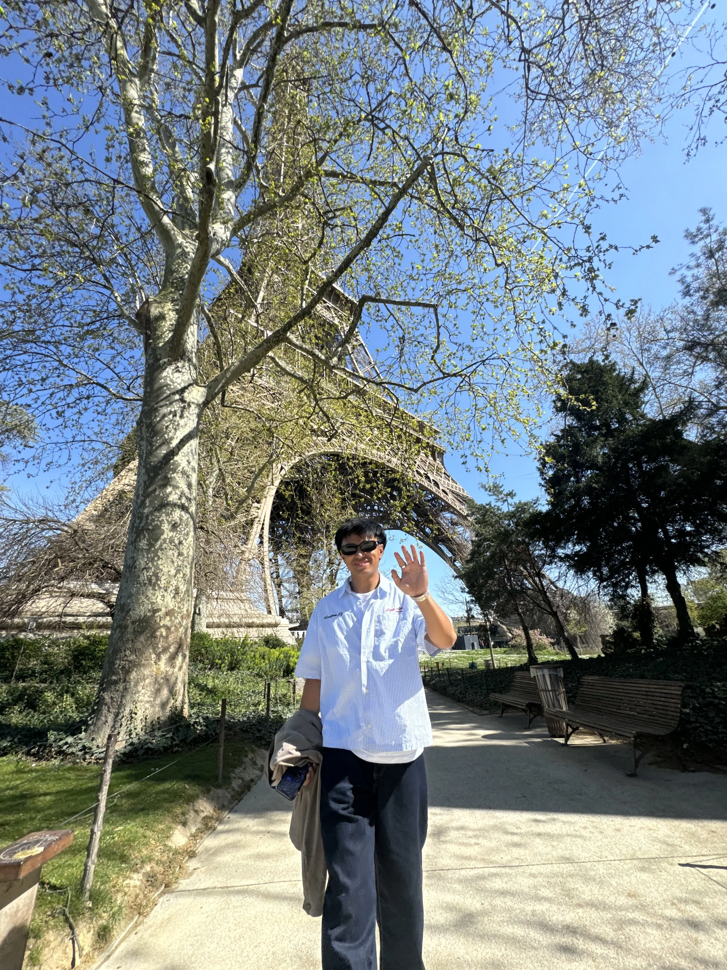 Person waving near the Eiffel Tower with a tree and clear blue sky.