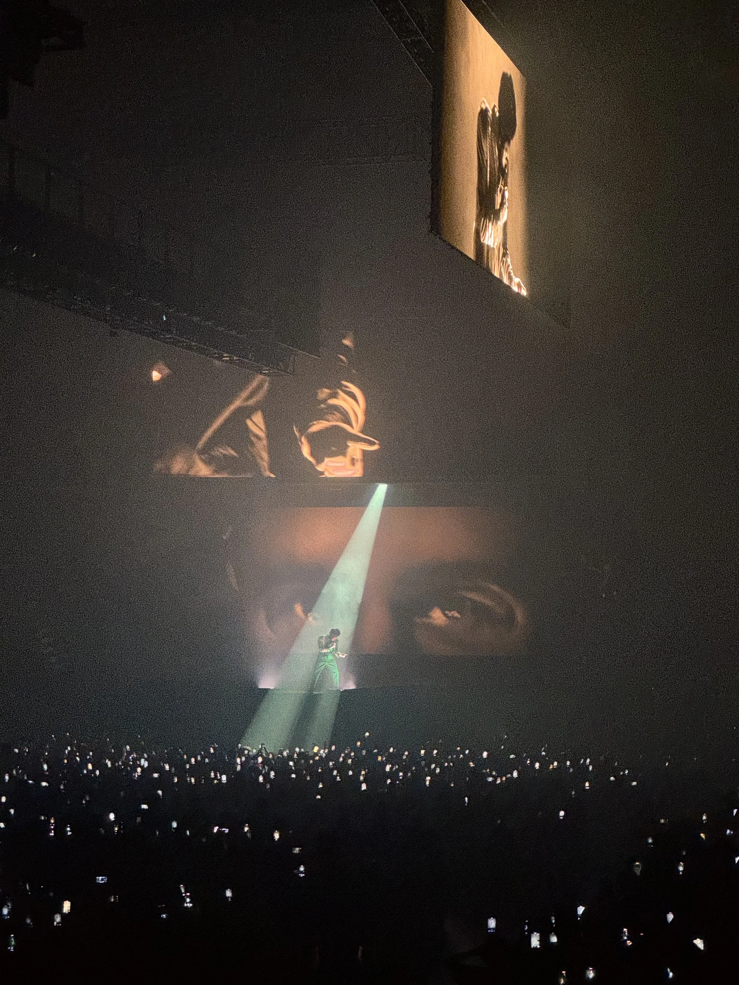 Concert scene with performer on stage under spotlight, large video screen showing close-up of eyes in background, audience members holding phones.